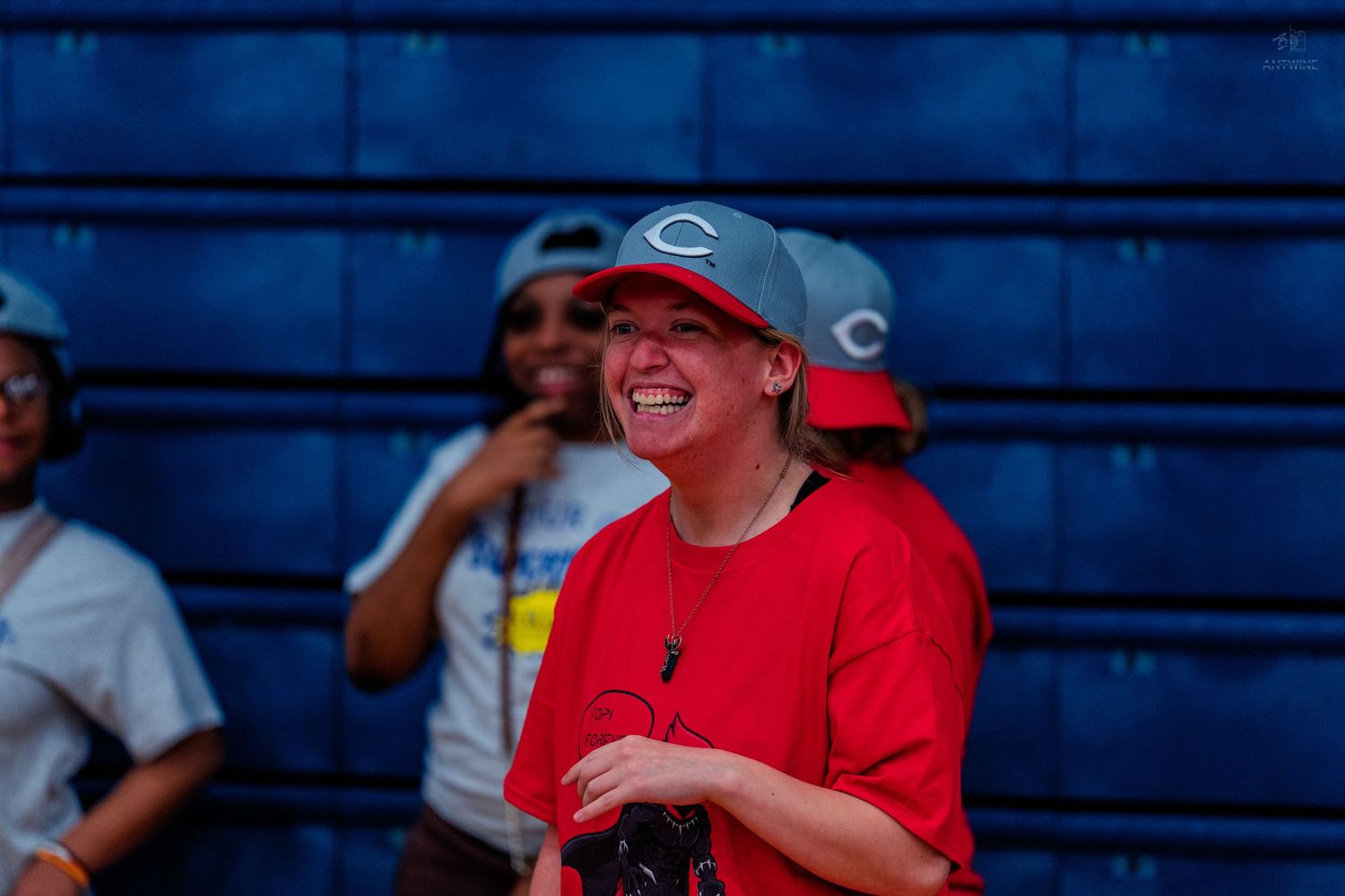 A smiling woman in a red shirt and Cincinnati Reds cap laughs joyfully outdoors near a blue wall.