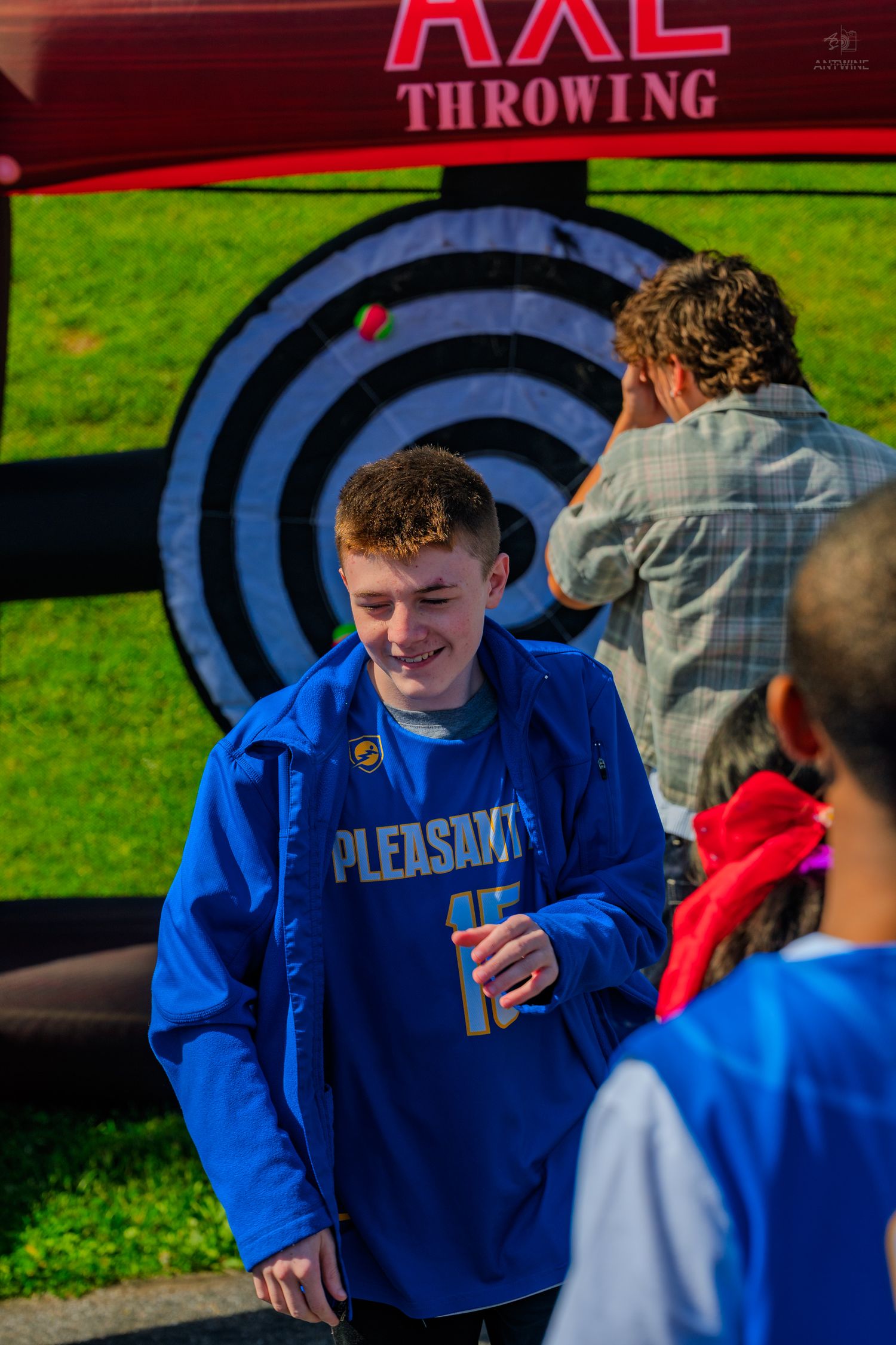 Smiling teen in blue Pleasants jersey at axe throwing activity outdoors.