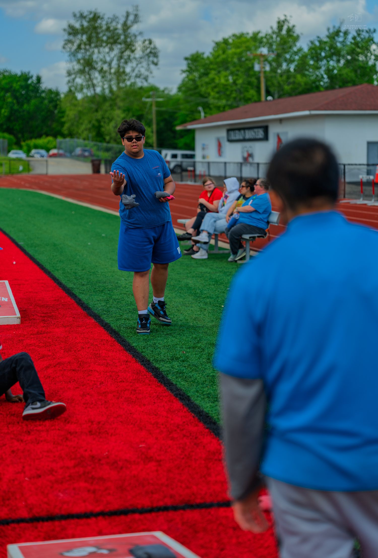 A coach in blue shorts instructs students on a red track at an outdoor athletic field with bleachers in the background.