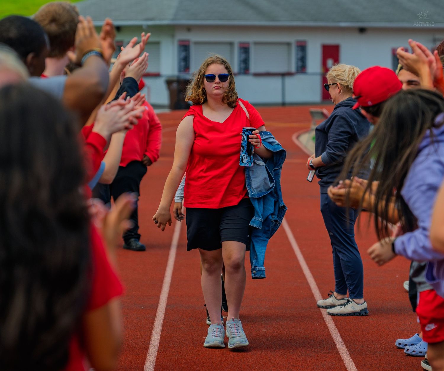 Woman in red top walks track flanked by cheering crowd during outdoor event.