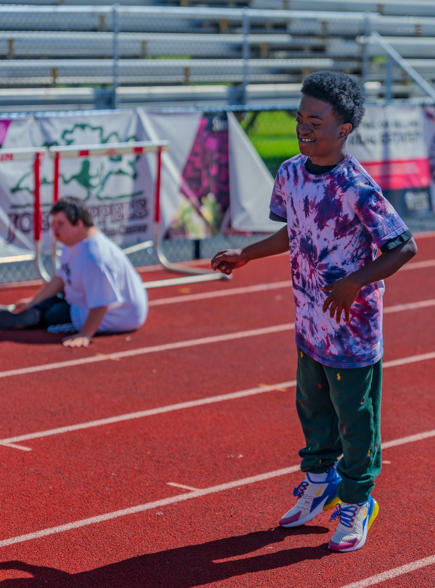 A smiling young boy in a tie-dye shirt and green pants stands on a red running track at a school stadium.