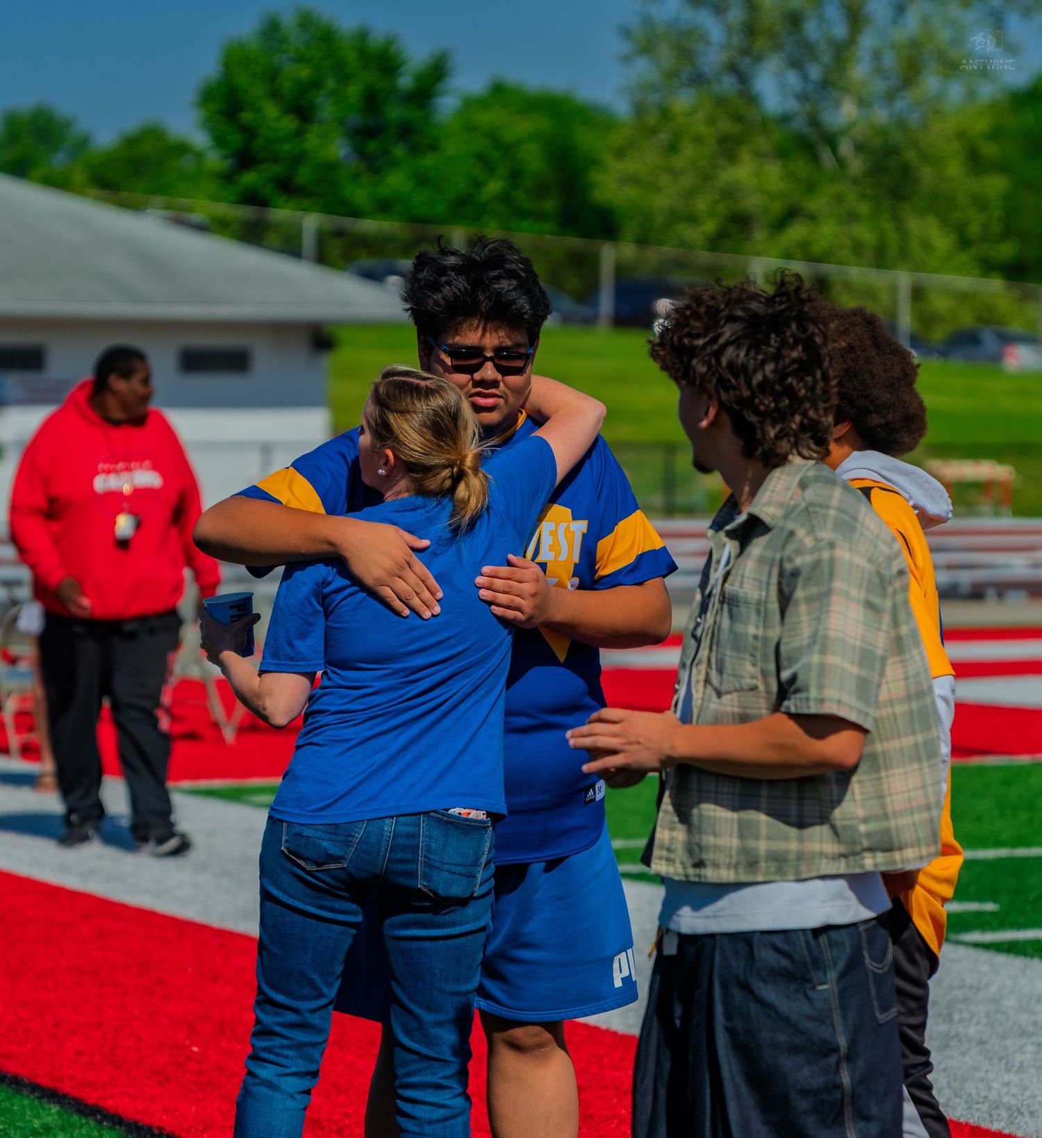 Students share an emotional hug on a red track field during an outdoor school sports event on a sunny day.