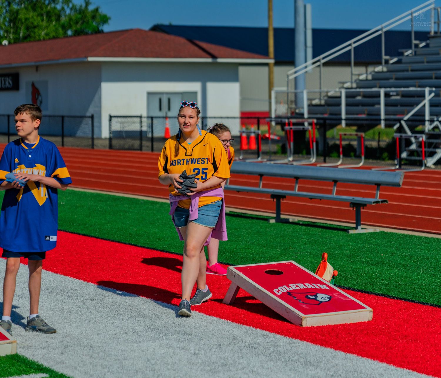 Students playing cornhole near a red track and bleachers at an outdoor school athletic event.