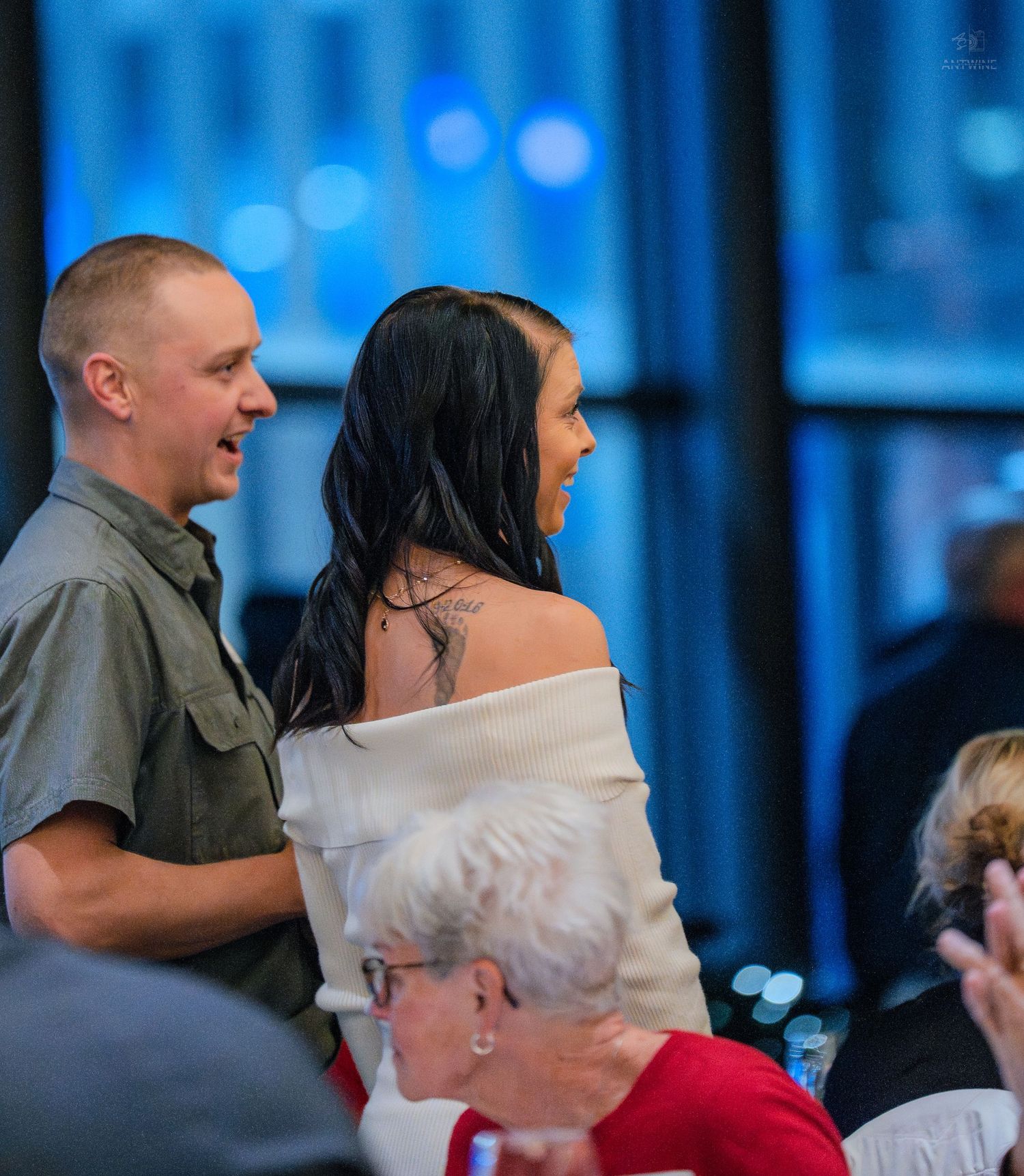 Couple standing together at an elegant indoor event, woman in off-shoulder white top with tattoo, man in olive shirt.