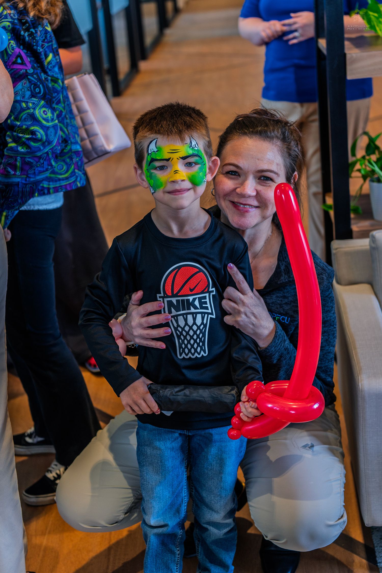 Smiling boy with green face paint and red balloon animal poses with a woman at a fun event.