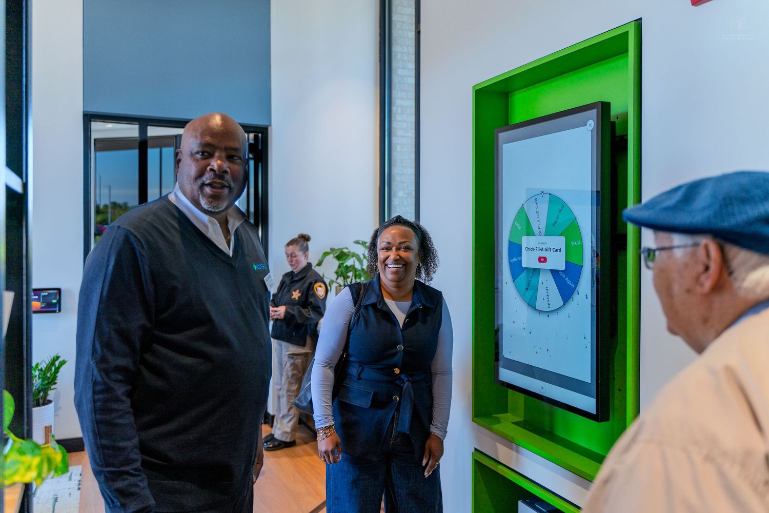 Two people smile at an interactive digital display screen mounted in a green frame inside a modern building.