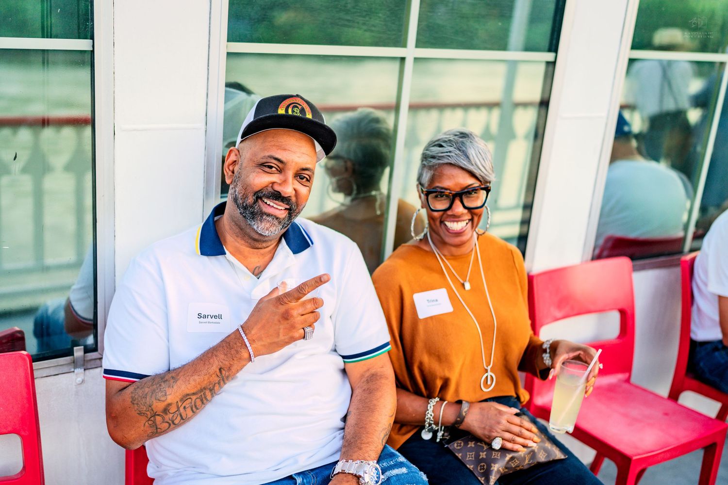 Two smiling friends pose together at an outdoor community event, man pointing at camera.