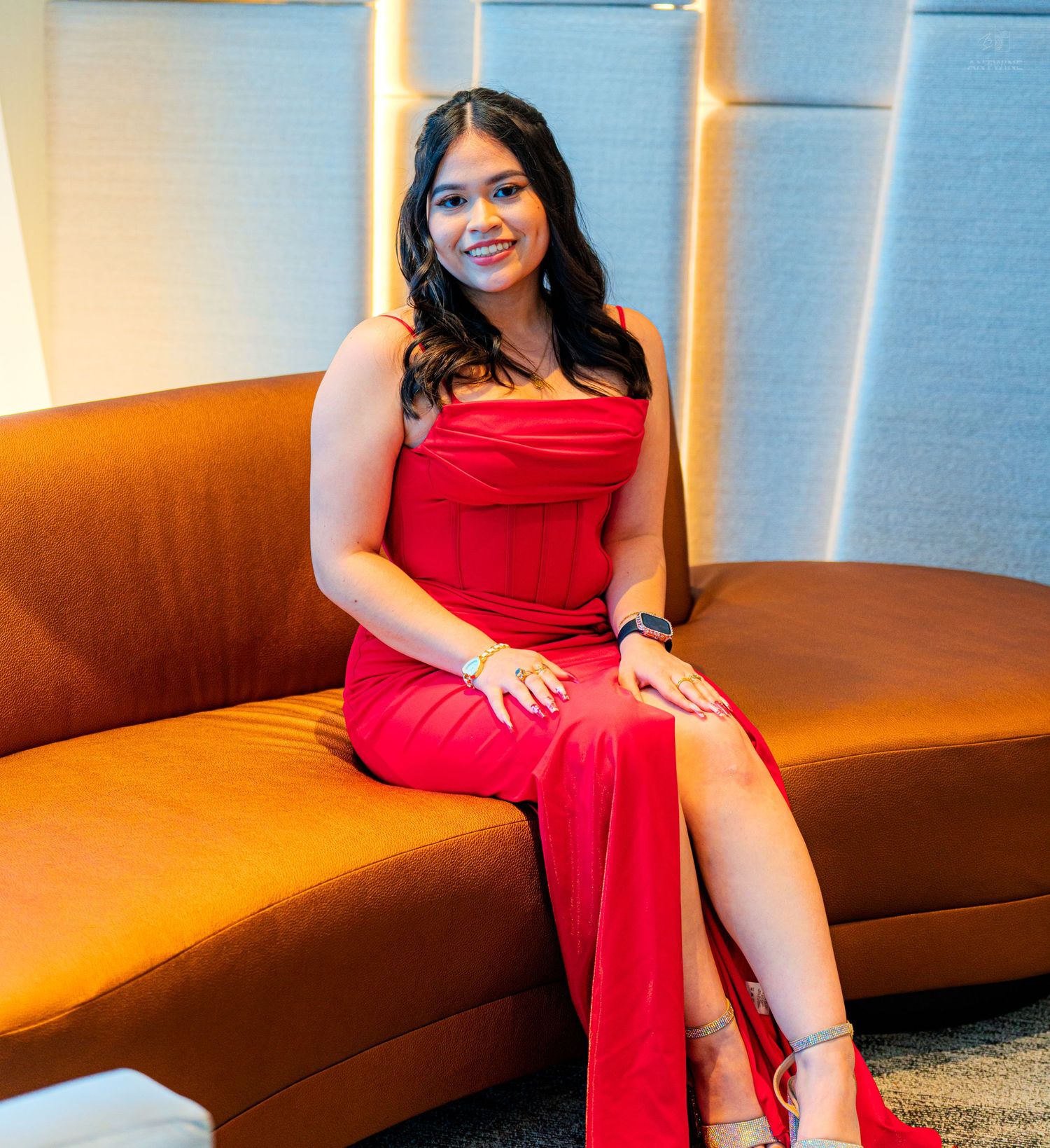 Woman in red strapless gown sitting elegantly on an orange curved sofa in a modern interior setting.