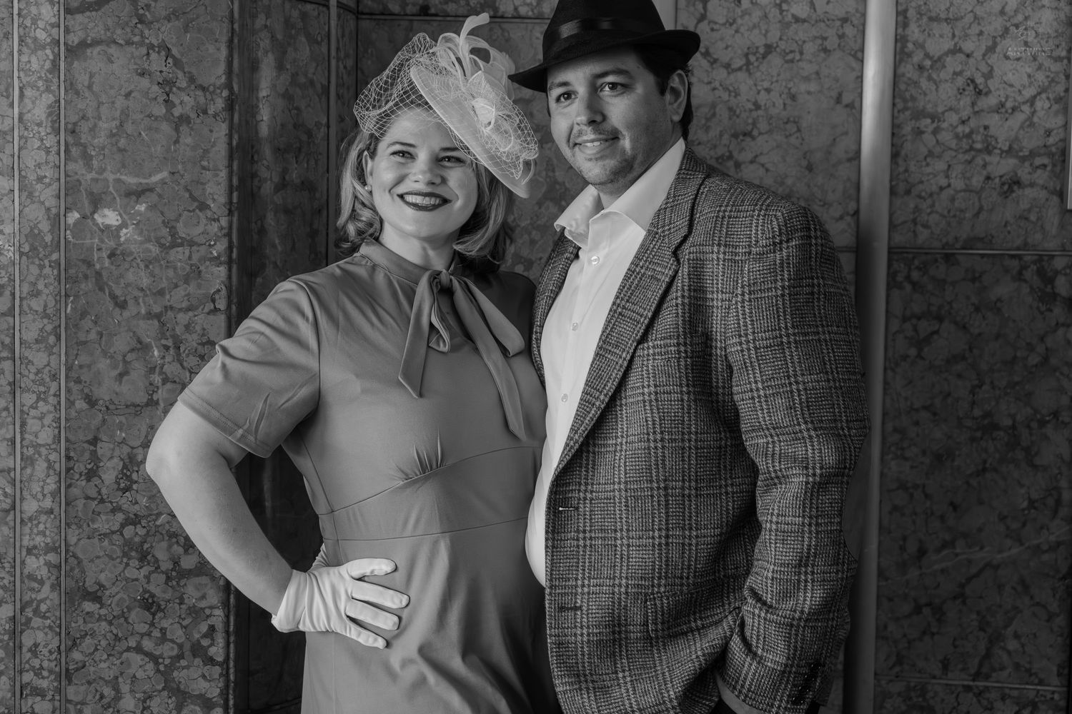 Black and white photo of an elegant couple in vintage attire, woman in dress with hat, man in plaid suit.