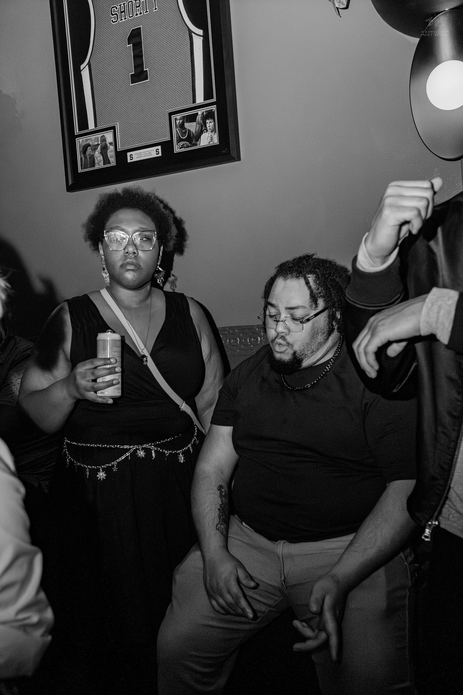 Black and white photo of two people at a party, woman holding a drink, man with dreadlocks, framed jersey on wall behind them.