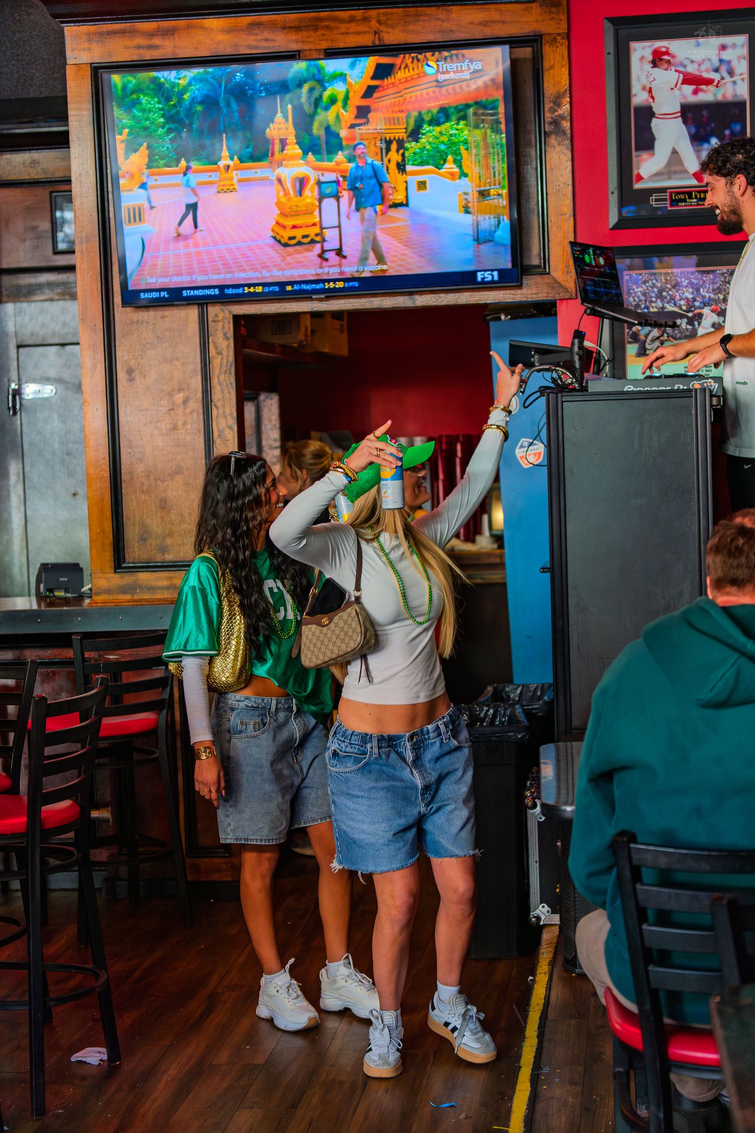 Two women playing an arcade game at a lively bar with a colorful video game on the TV screen.