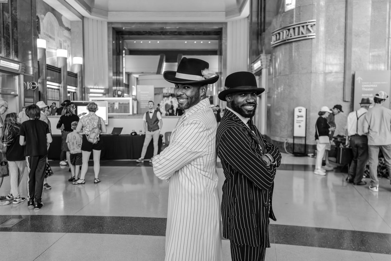 Two men in pinstripe suits and fedoras pose back-to-back in a grand hall, black and white photo.