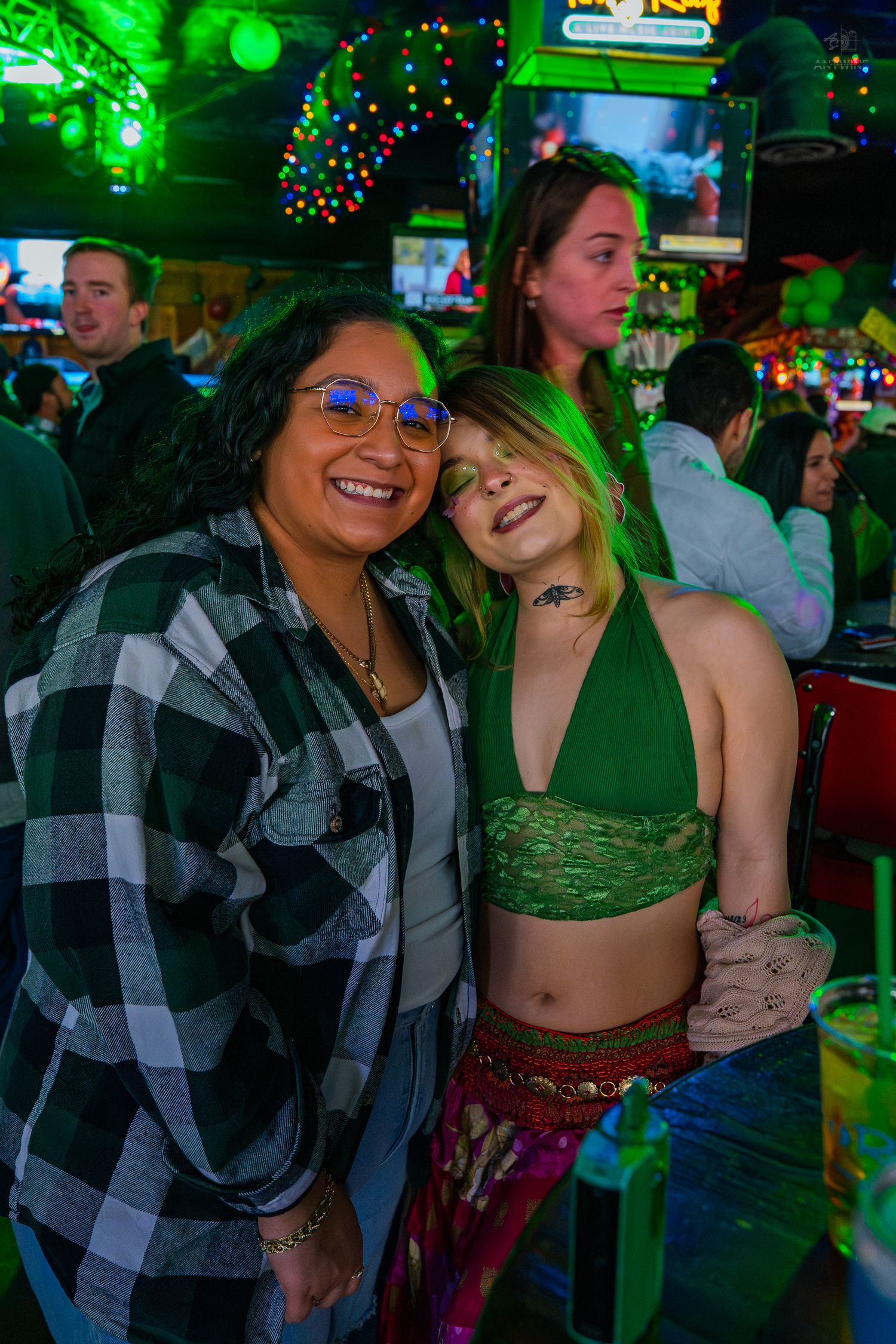 Two women smiling and posing together at a festive bar with green lights during St. Patrick's Day celebration.