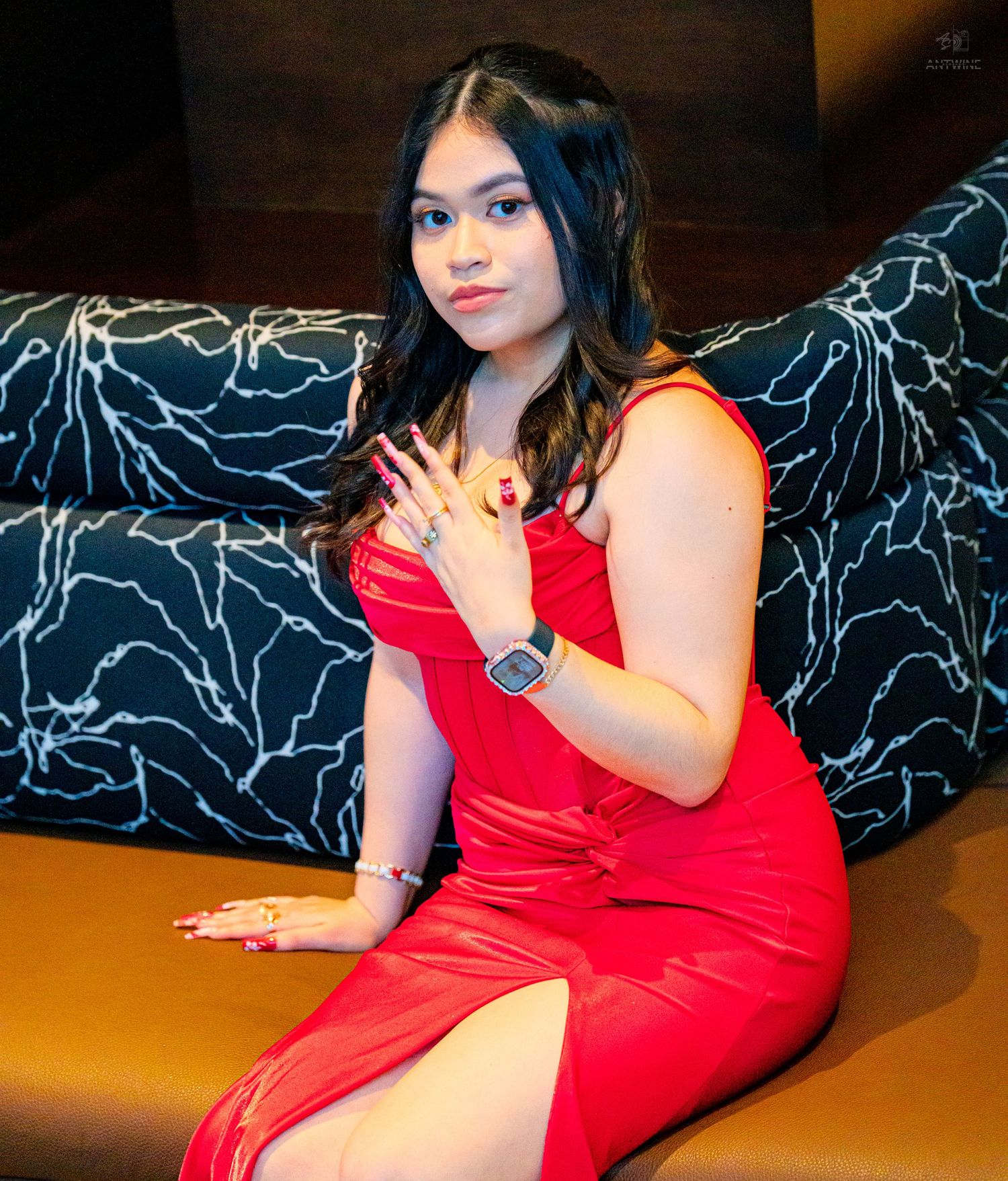 Young woman in red slit dress sitting on couch wearing a square watch.