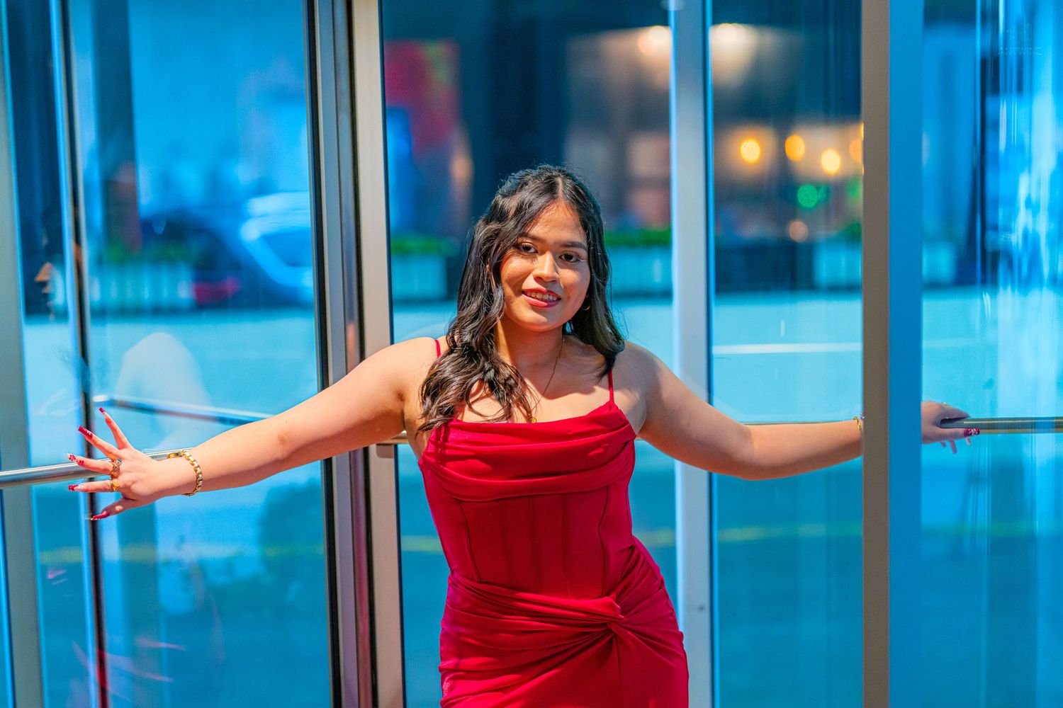 Smiling woman in red dress poses with arms spread at glass revolving doors.