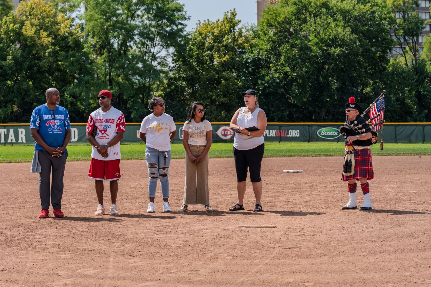 Group of six people and a bagpiper standing on a softball diamond during an outdoor ceremonial event.