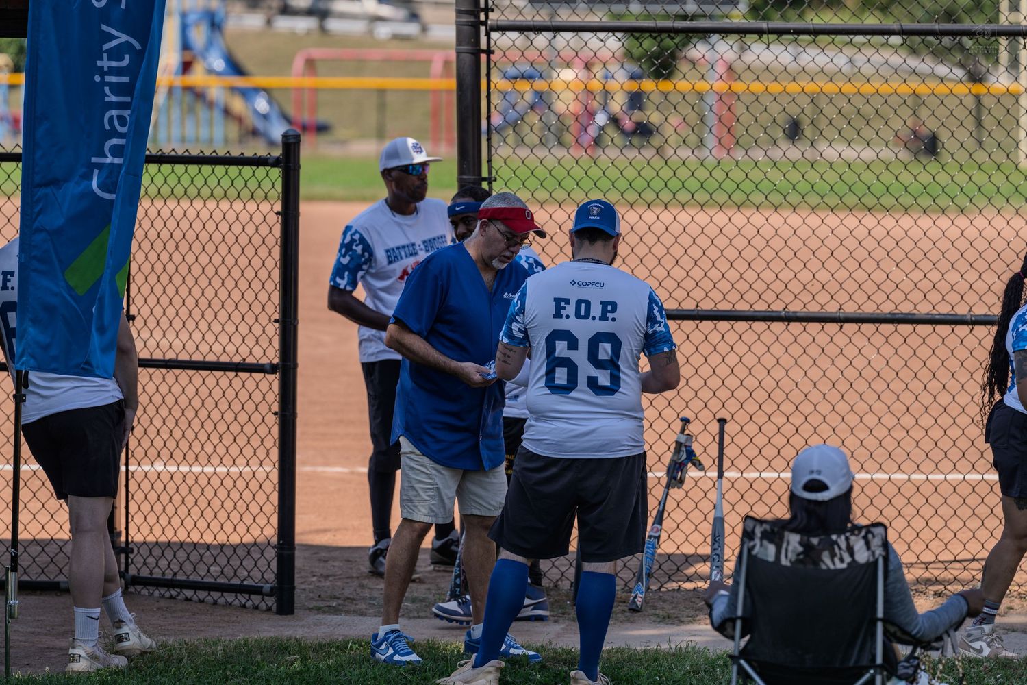 Men in blue FOP 69 jerseys interact at a charity softball game on a sunny day near a chain-link fence.
