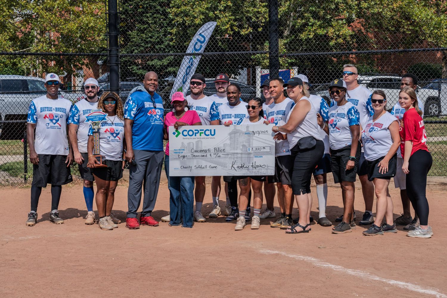 Large group poses with an oversized COPFCU charity check at a softball fundraiser event.