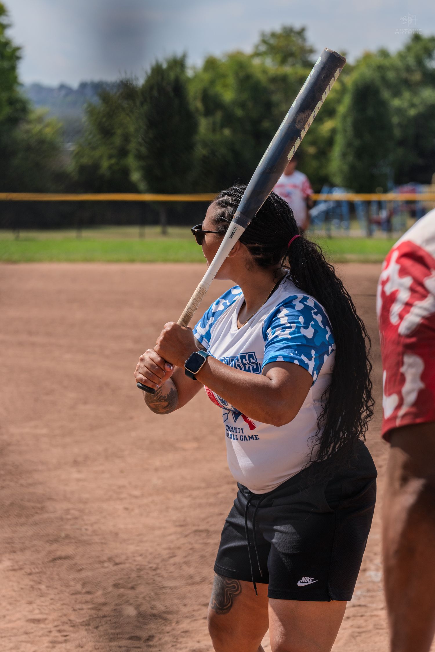 Woman in blue softball uniform holds bat ready at plate during outdoor charity softball game.