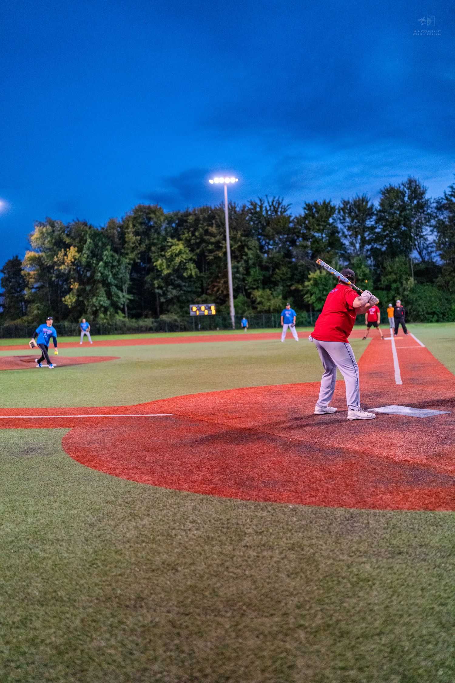 A batter in a red shirt prepares to swing during a night baseball game under bright field lights with players positioned in the outfield.