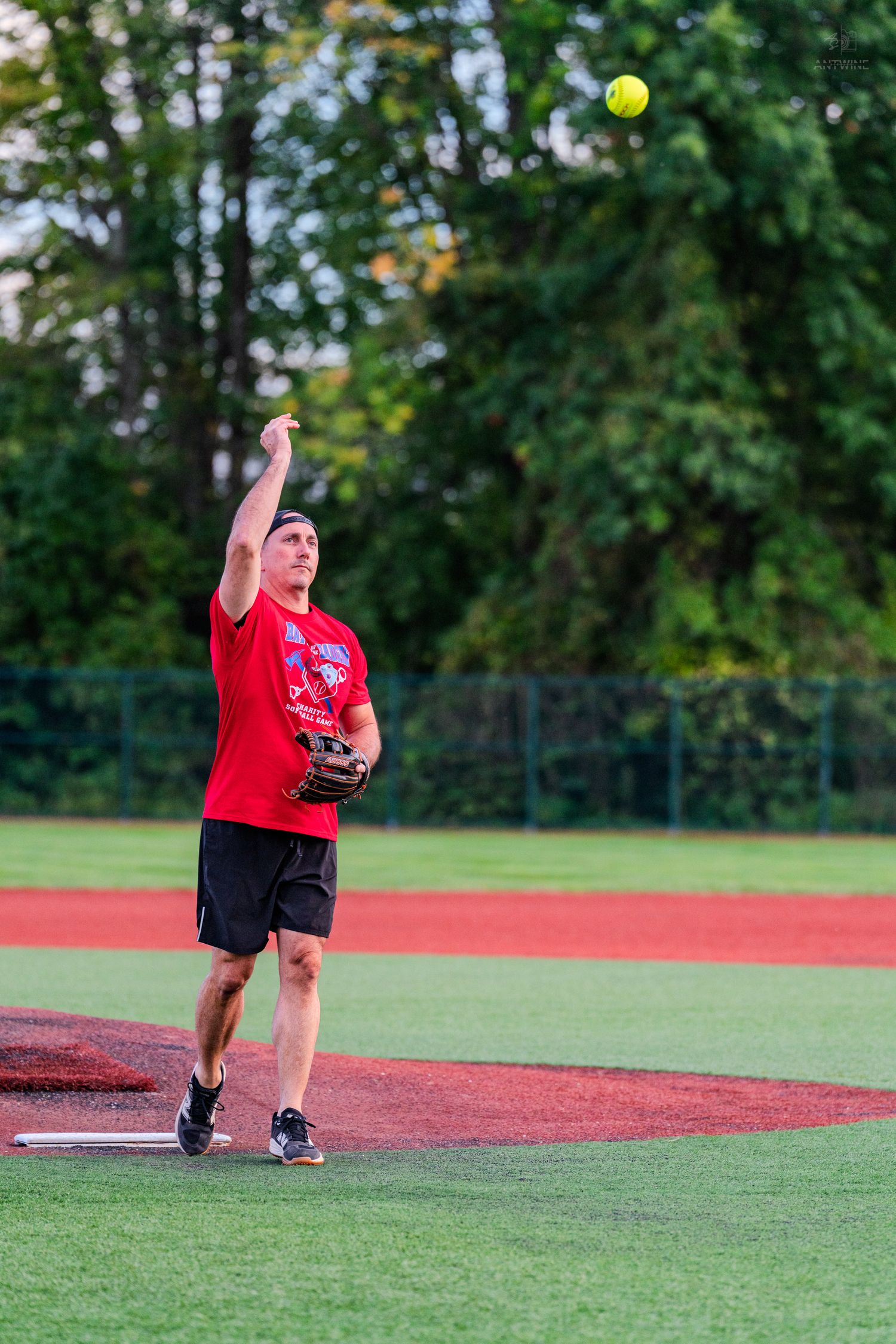 A pitcher in a red shirt and black shorts releases a yellow softball from the mound outdoors.