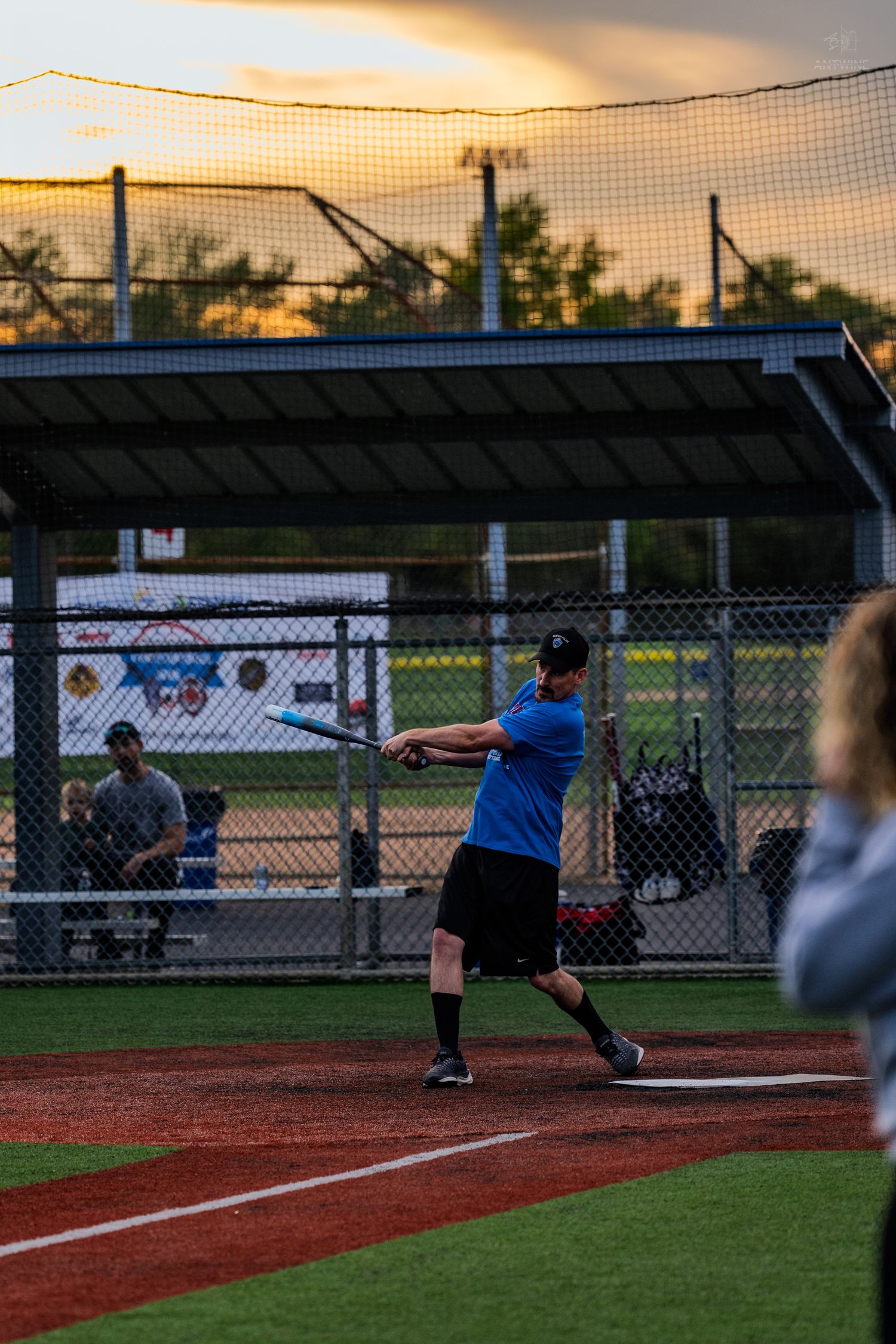 Softball batter in blue jersey swings at pitch during sunset recreational league game at outdoor ballpark.