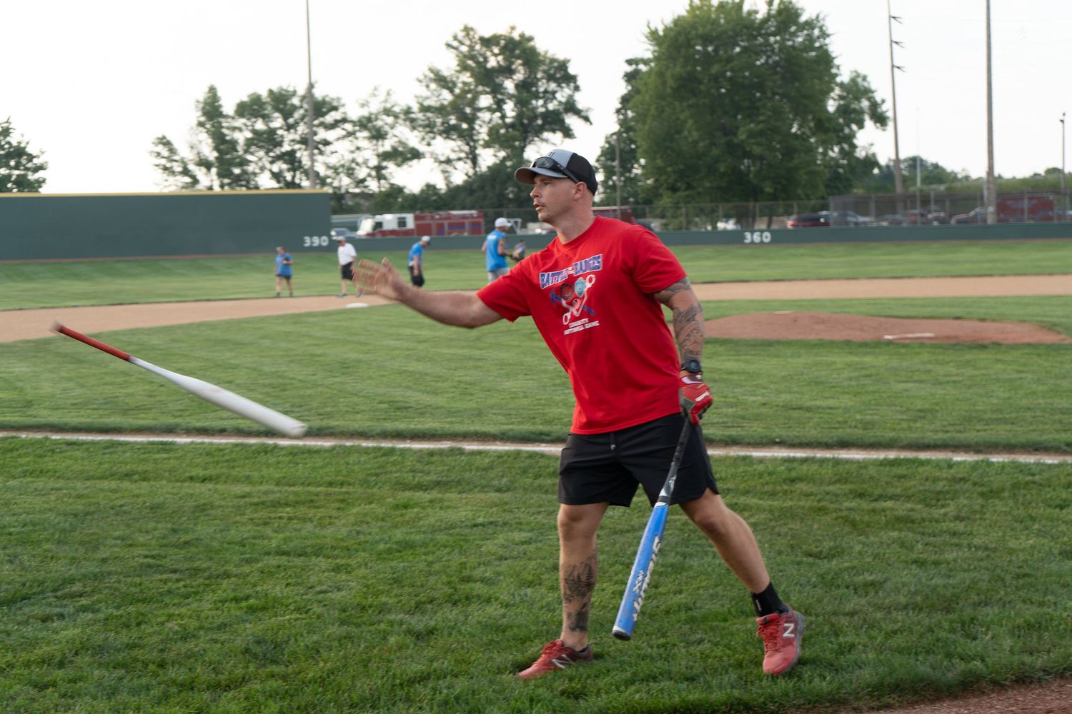 Man in red shirt throwing a baseball bat on an outdoor baseball field during evening practice.