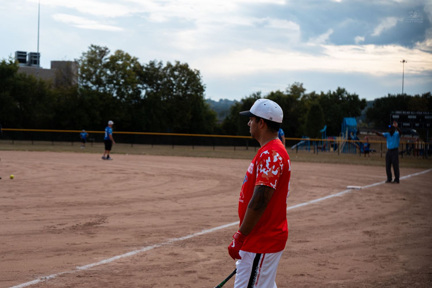 Baseball player in red jersey holding a bat on a dirt field at dusk during a game.