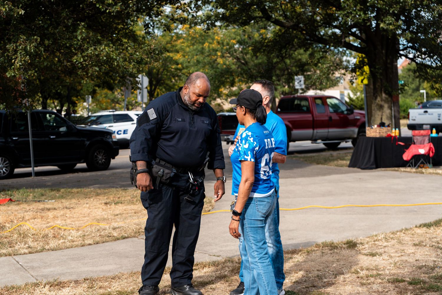 A police officer speaks with a civilian outdoors under trees at a community event on a sunny day.
