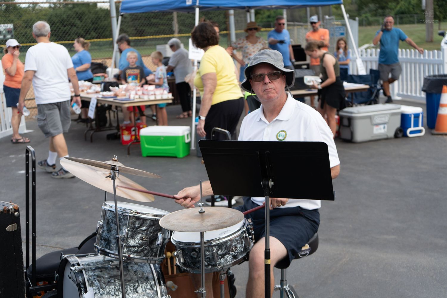 Man playing drums outdoors at a community event with people browsing tables in the background.