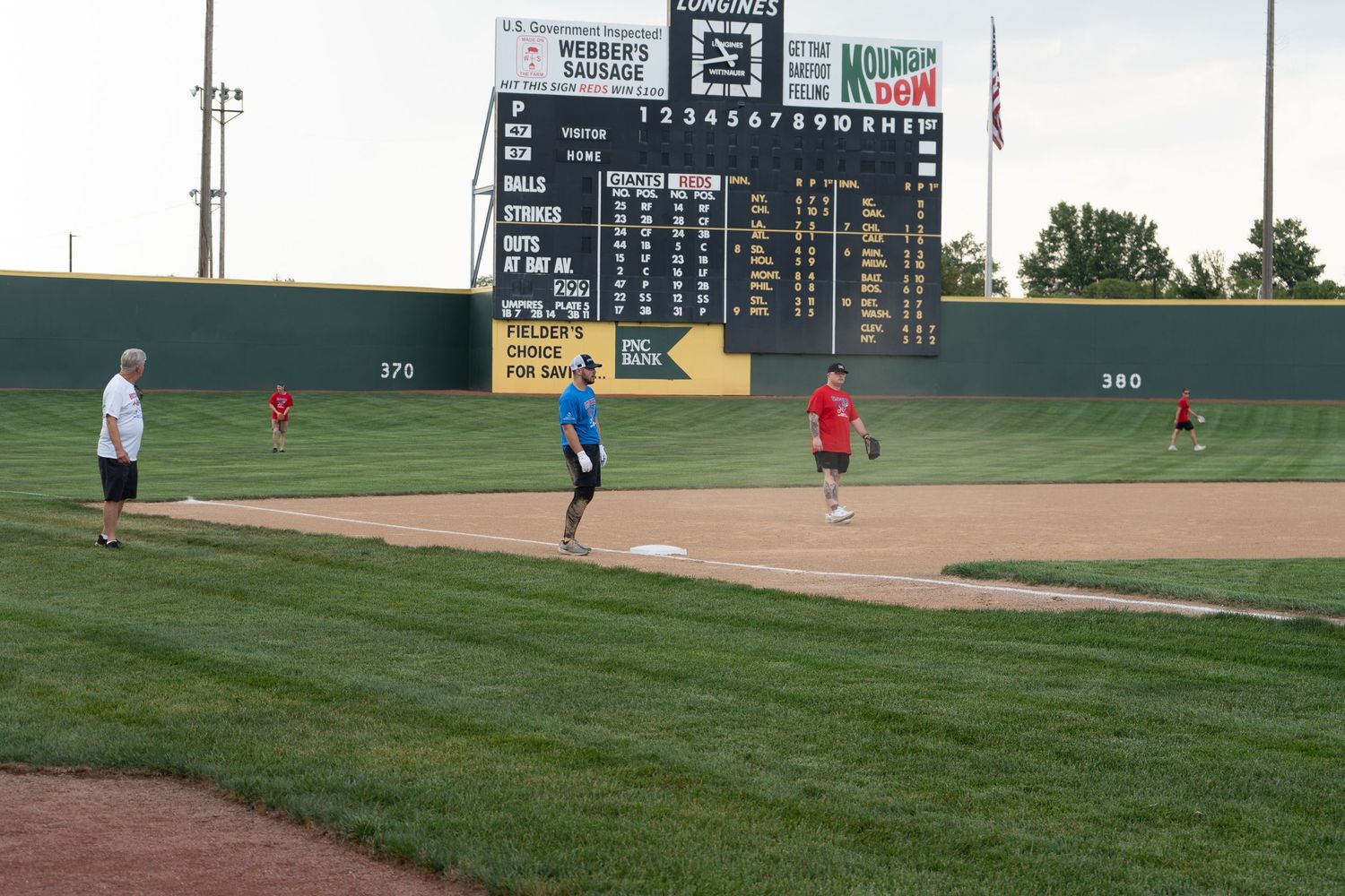 Players enjoy a softball game on a professional field with a large scoreboard and green outfield walls visible.