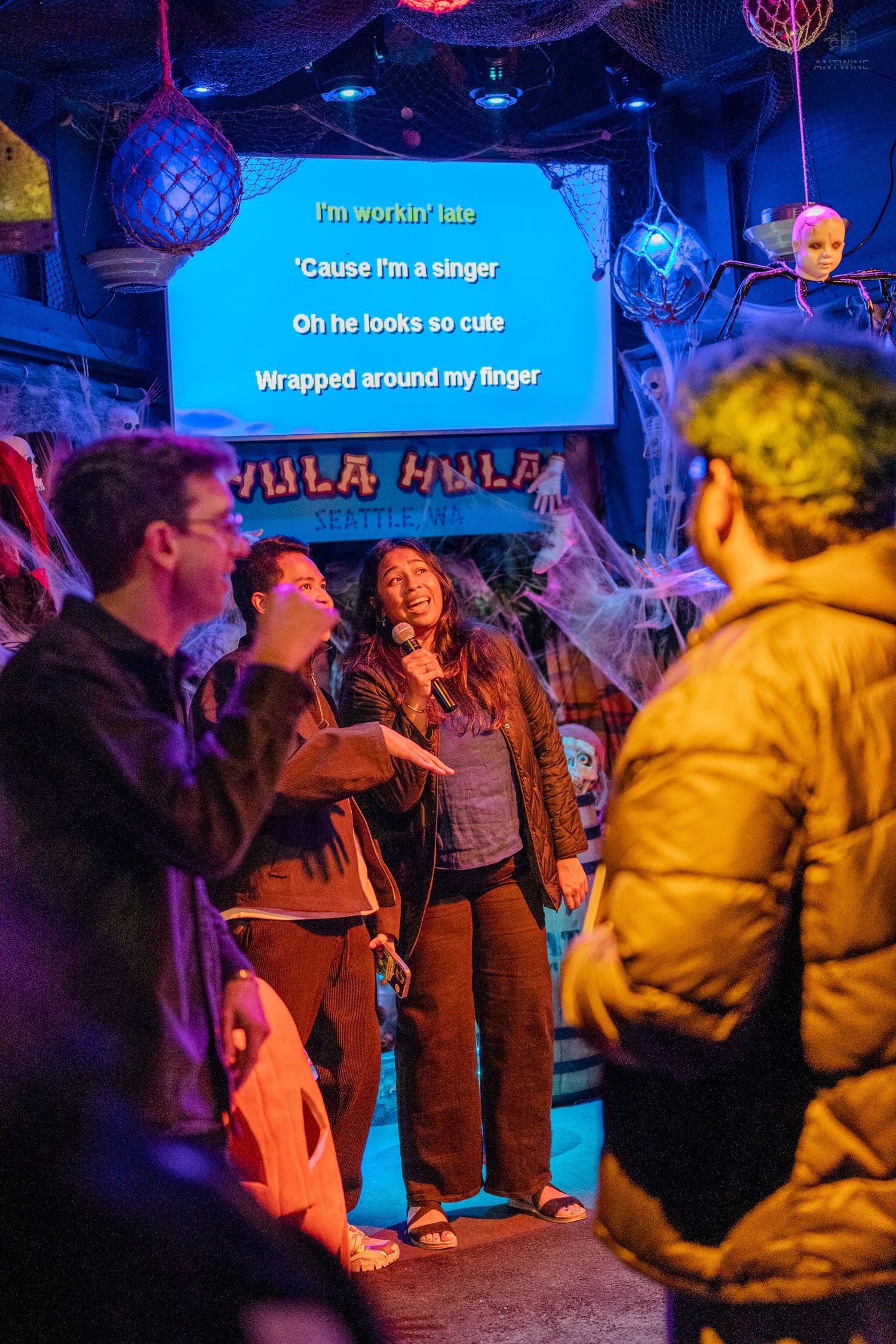 Woman sings karaoke at Hula Hula bar decorated with Halloween cobwebs while crowd watches lyrics screen.