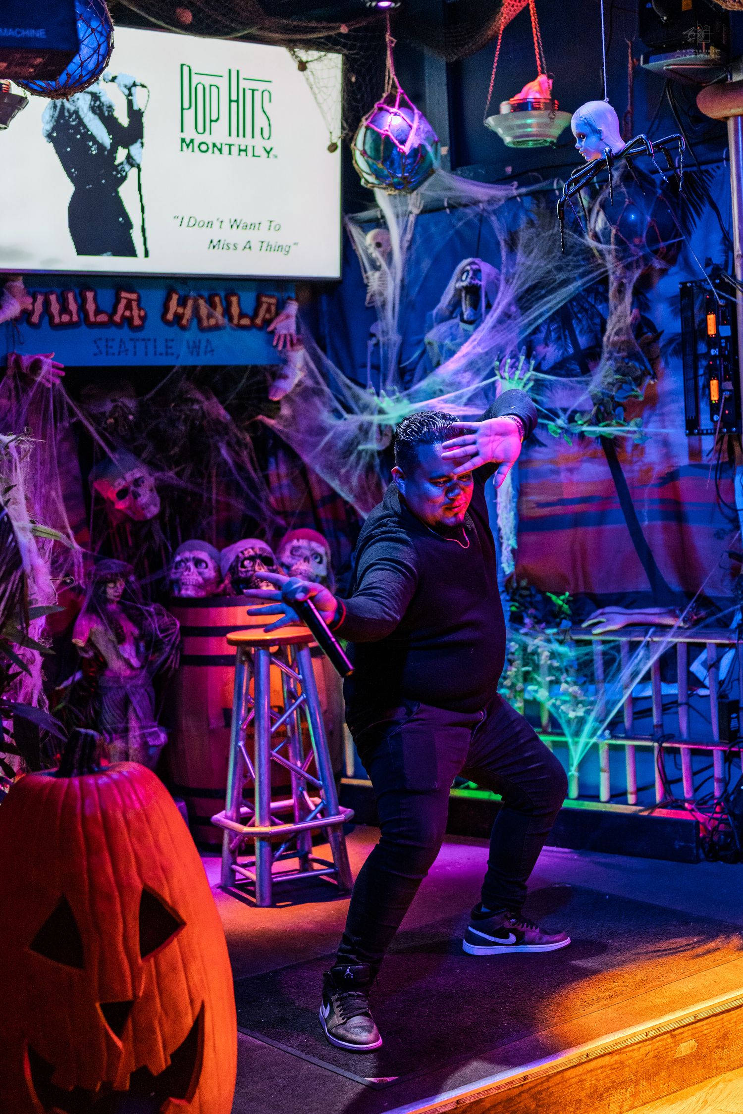 Performer dancing on Halloween-decorated stage with spooky skeletons, cobwebs, jack-o-lantern, and Pop Hits Monthly display.