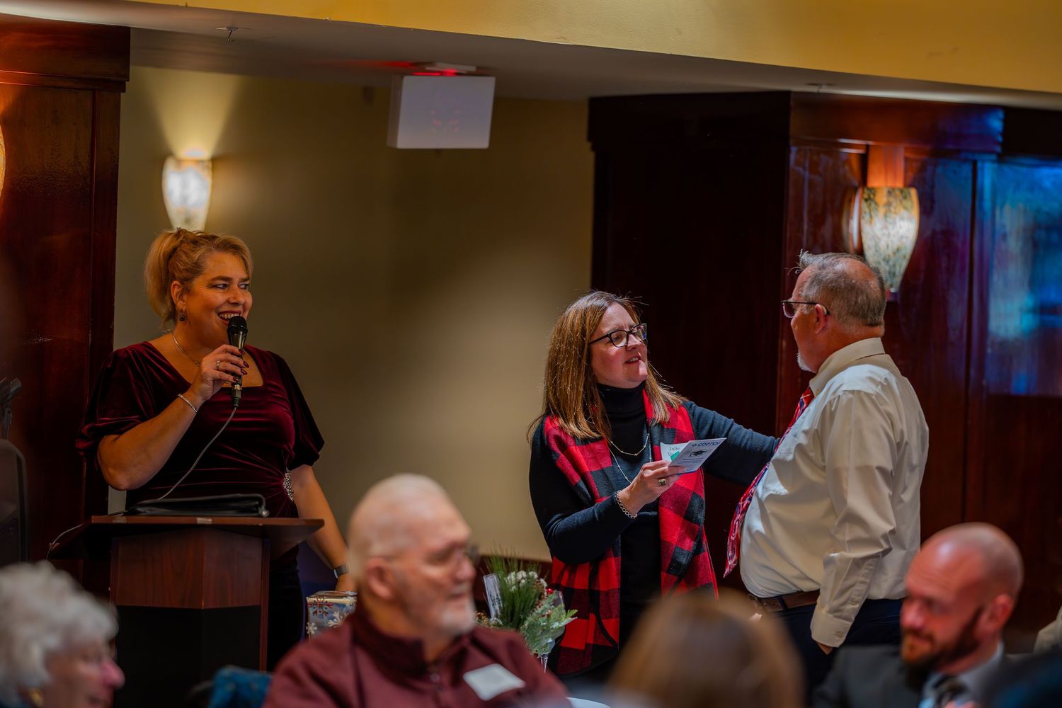 Three people engaged in conversation at an indoor event, with a woman speaking into a microphone at a podium.