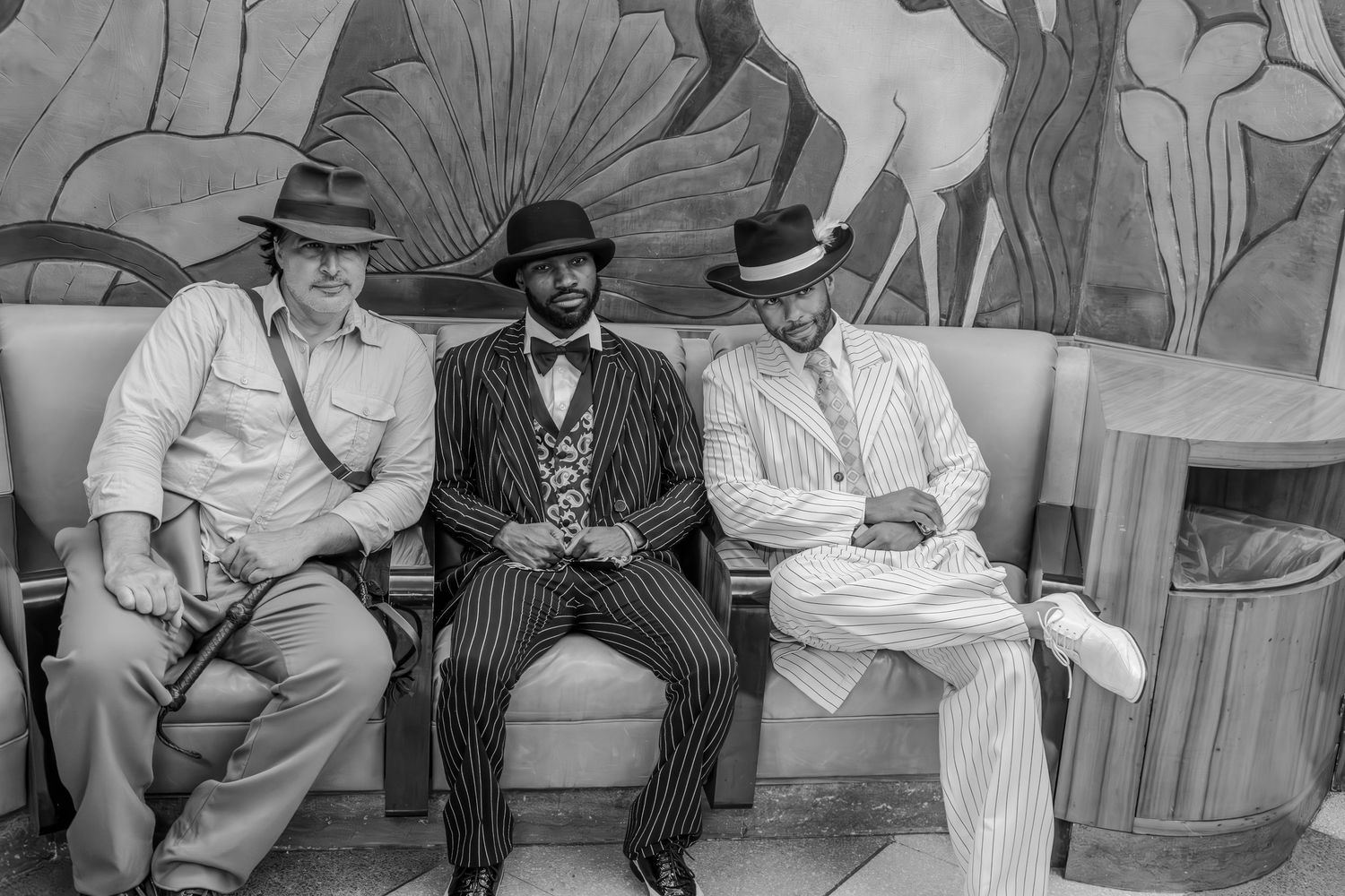 Three men in vintage suits and hats sit on a bench before an Art Deco mural in a black and white photo.