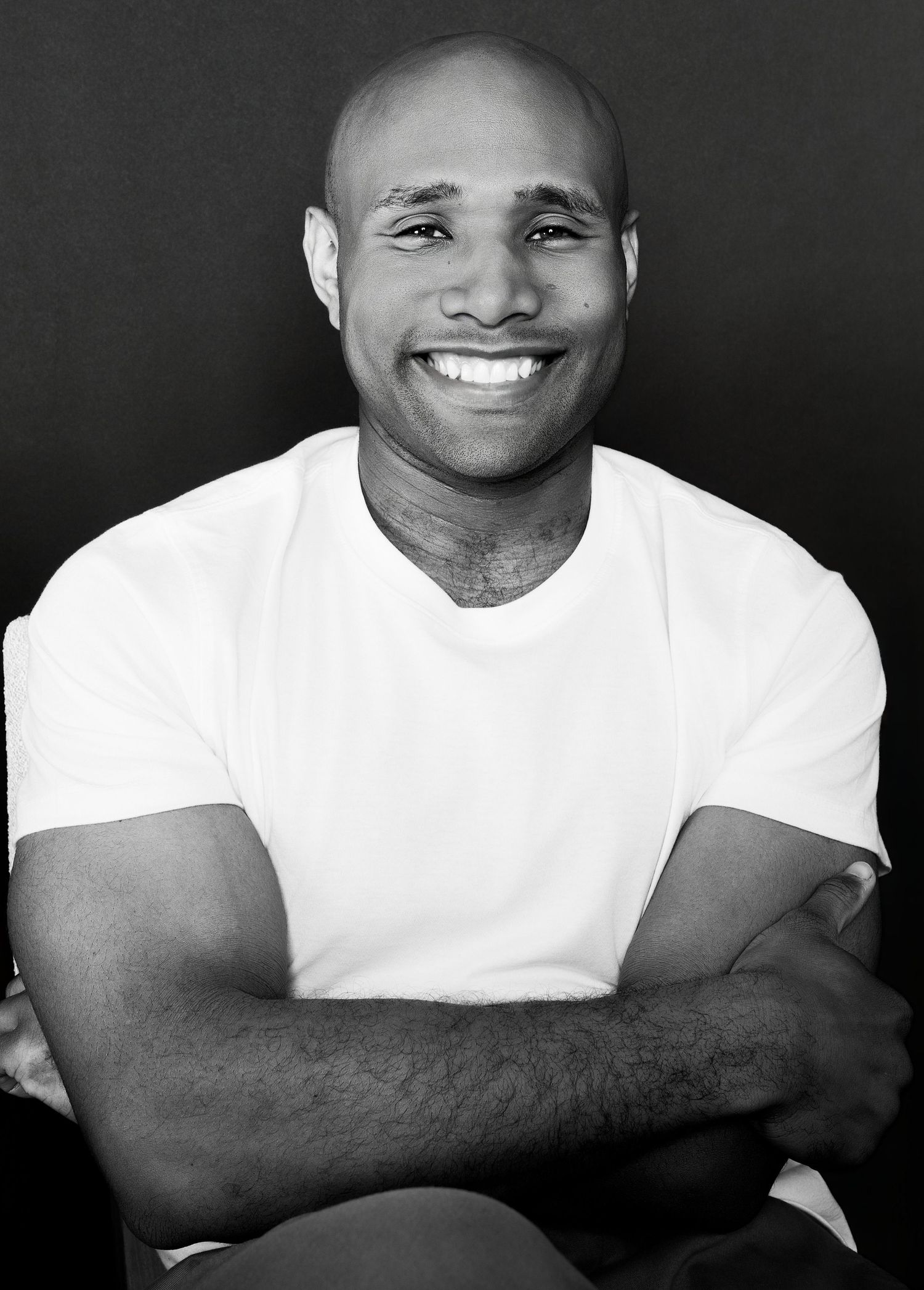 Smiling bald Black man in white t-shirt with arms crossed, black and white portrait photo against dark background.