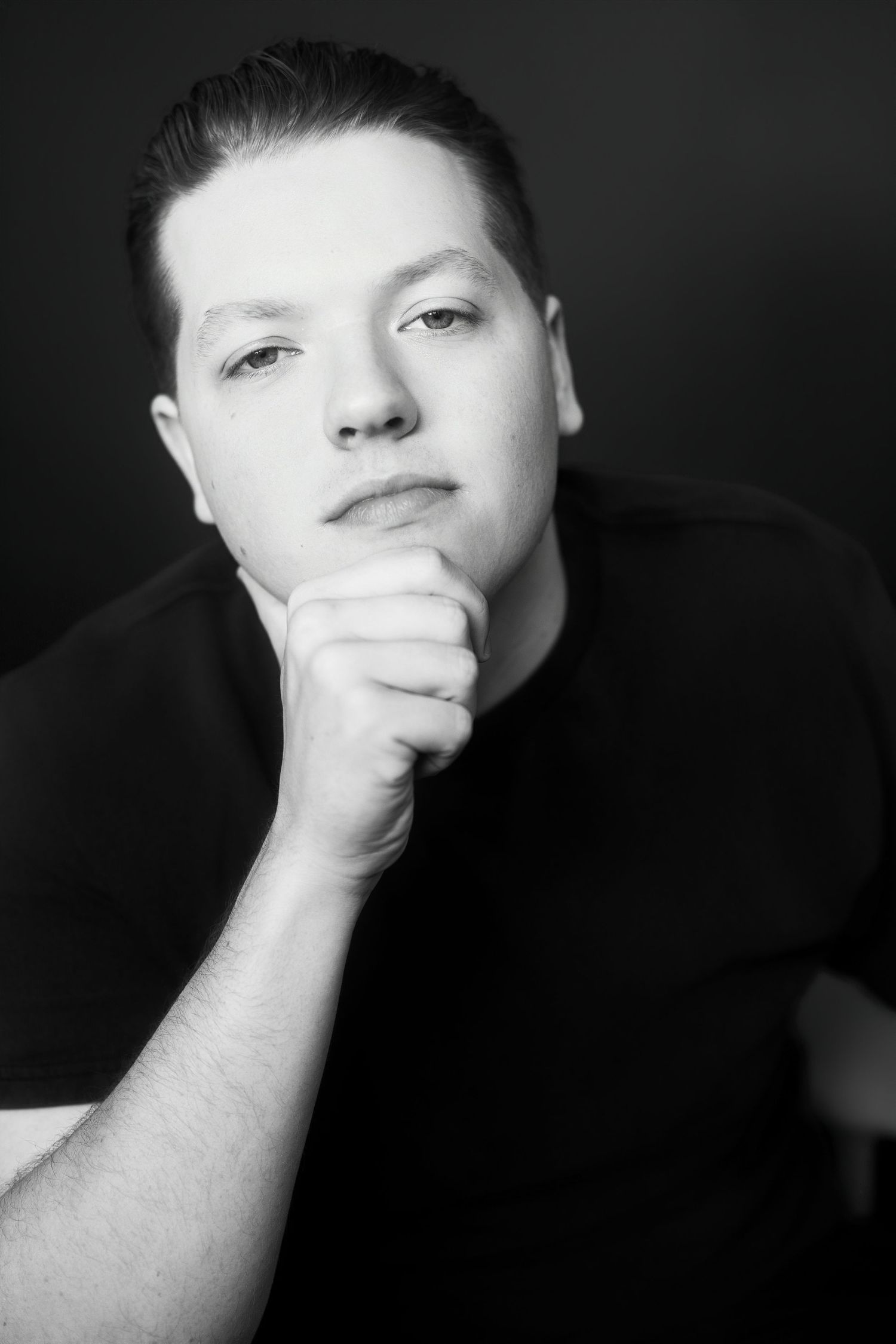 Black and white studio portrait of young man with fist under chin against dark background.