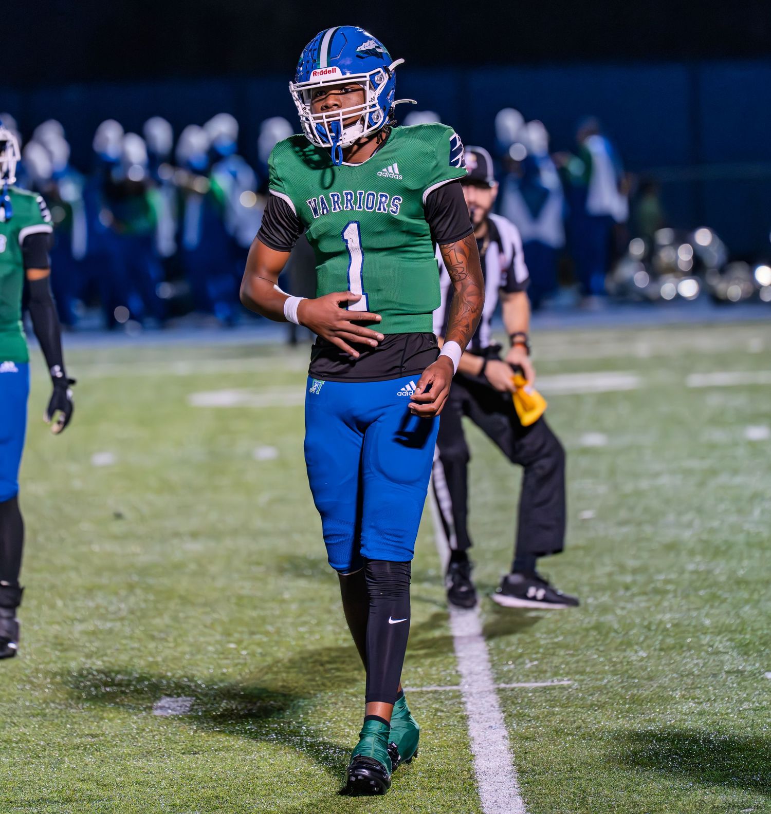 A football player wearing number 1 in a green Warriors uniform walks on the field during a night game.