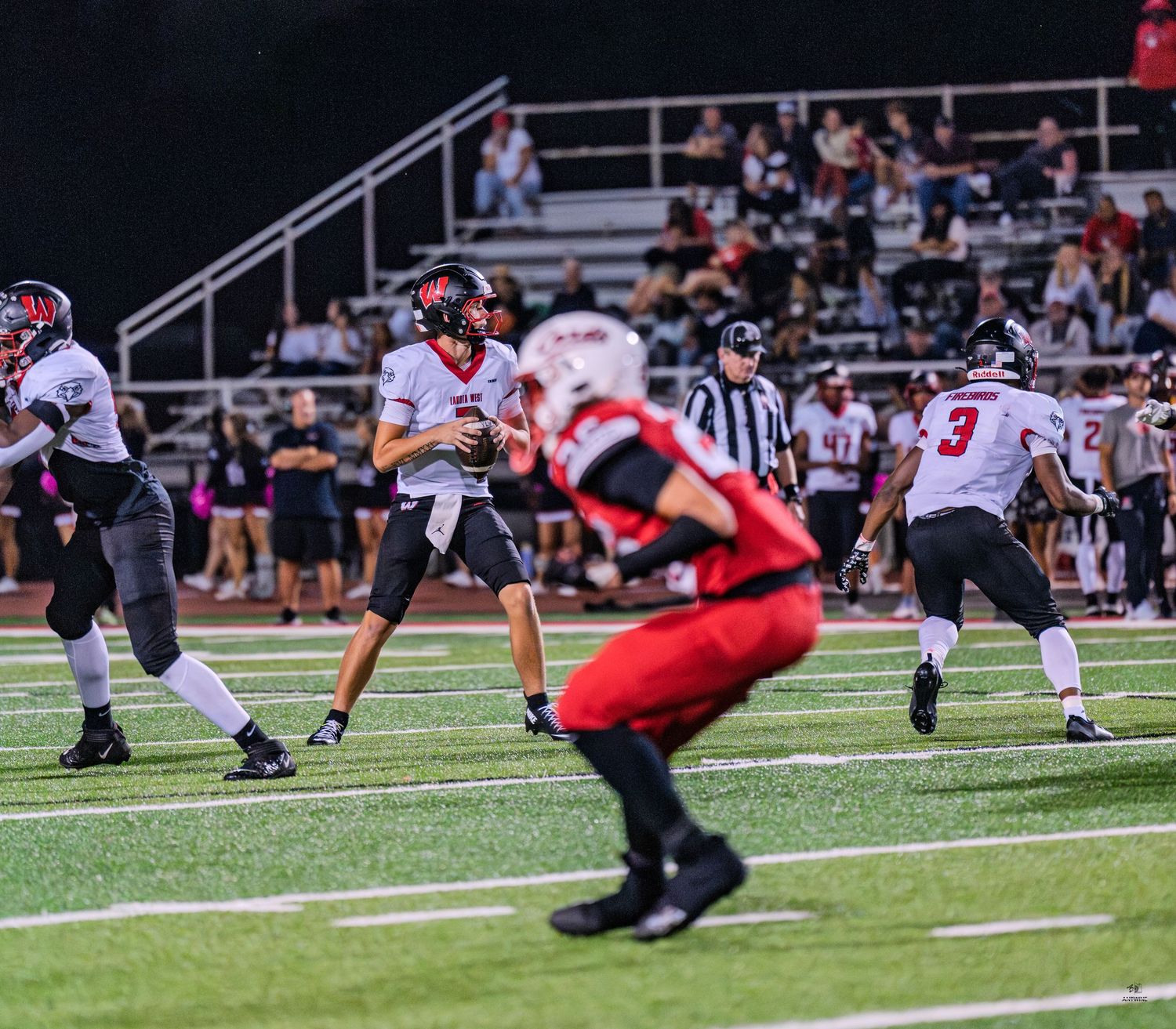 A quarterback in dark uniform drops back to pass while Colerain defenders in red rush during a night game.