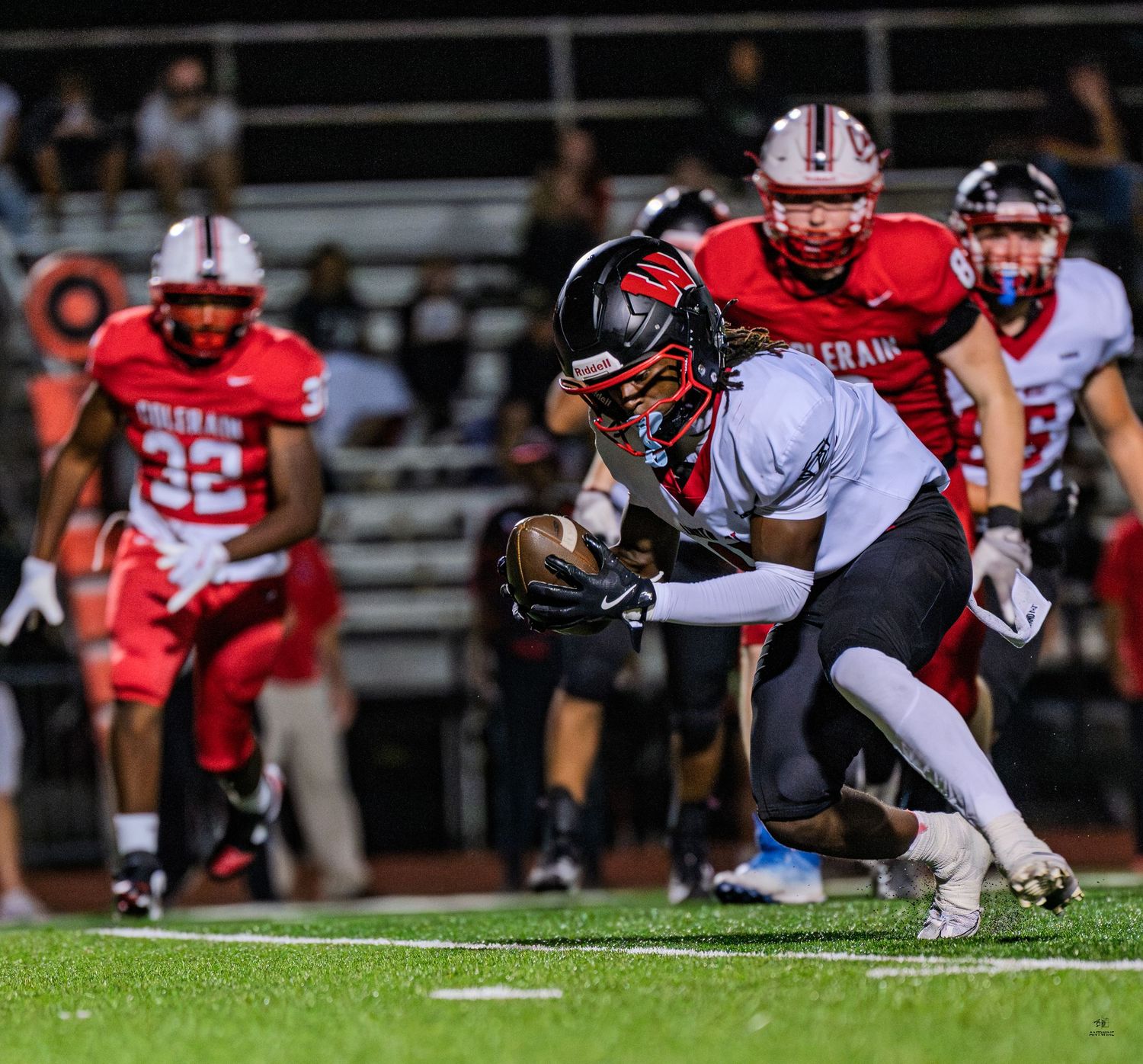 Football player in white uniform catches ball while defenders in red pursue during night game.