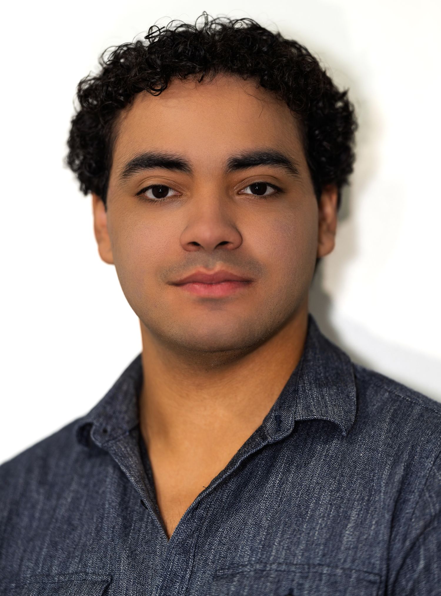 Young man with curly black hair in a blue denim shirt posing for a professional color headshot.