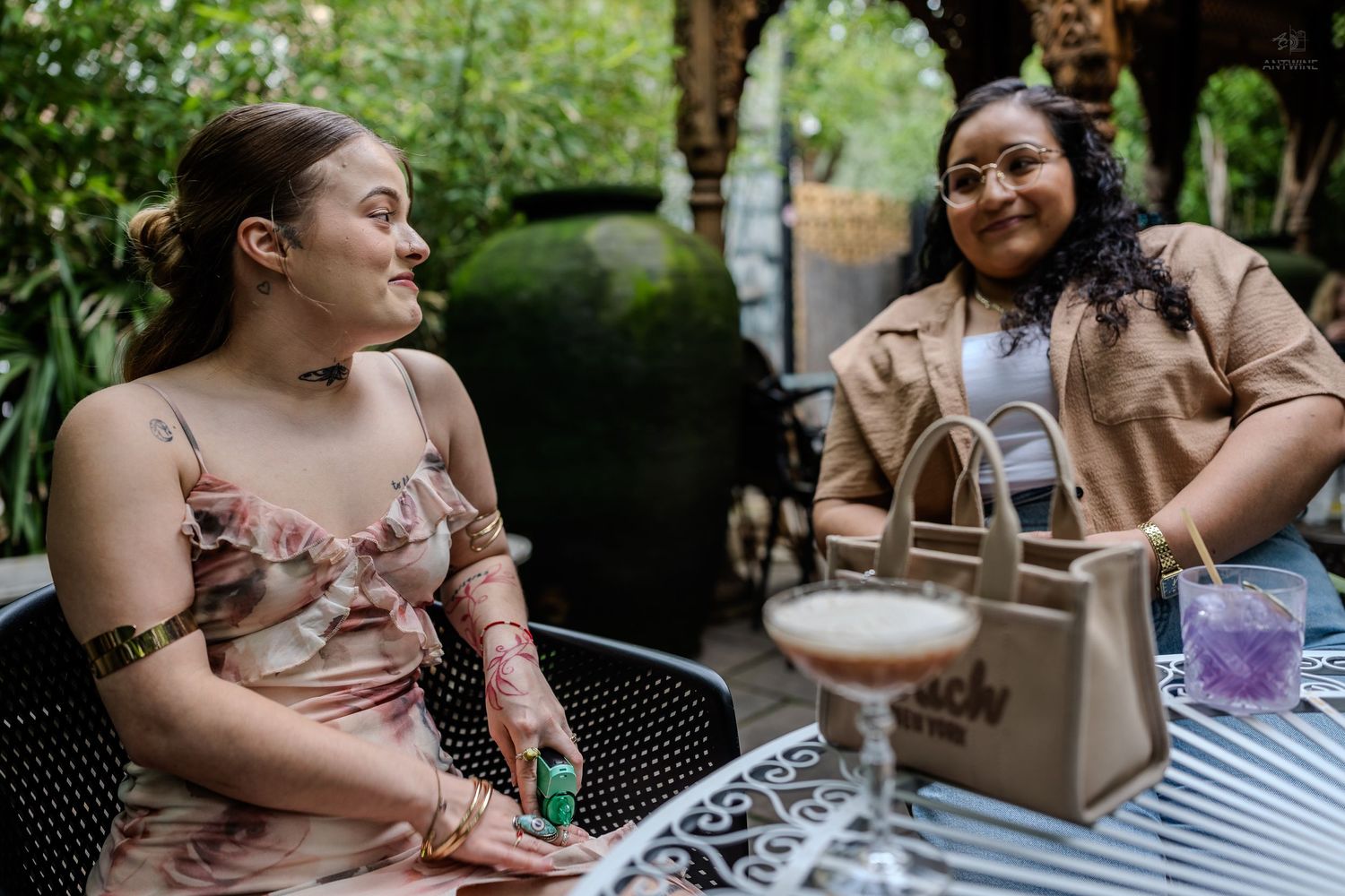 Two women laughing and enjoying cocktails at outdoor patio table surrounded by lush greenery.