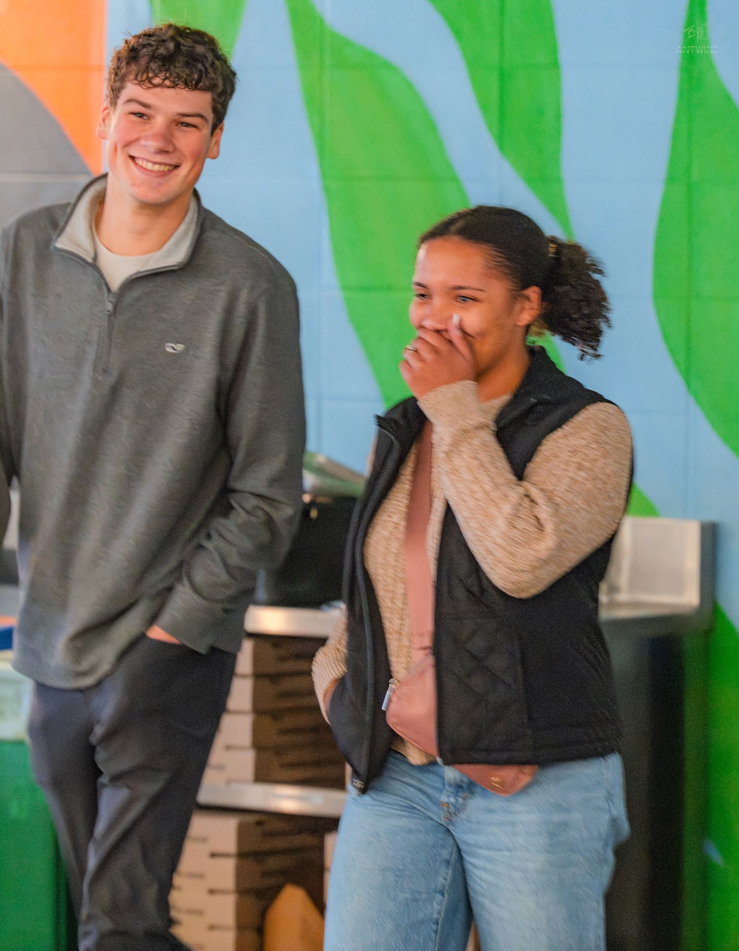A smiling young man in a gray pullover stands beside a laughing woman in a black vest near a colorful mural.
