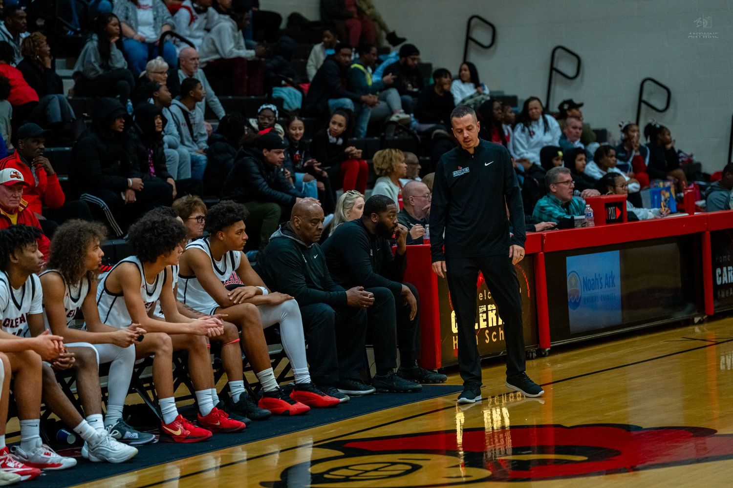 High school basketball coach stands on sideline watching players during a packed gym game.