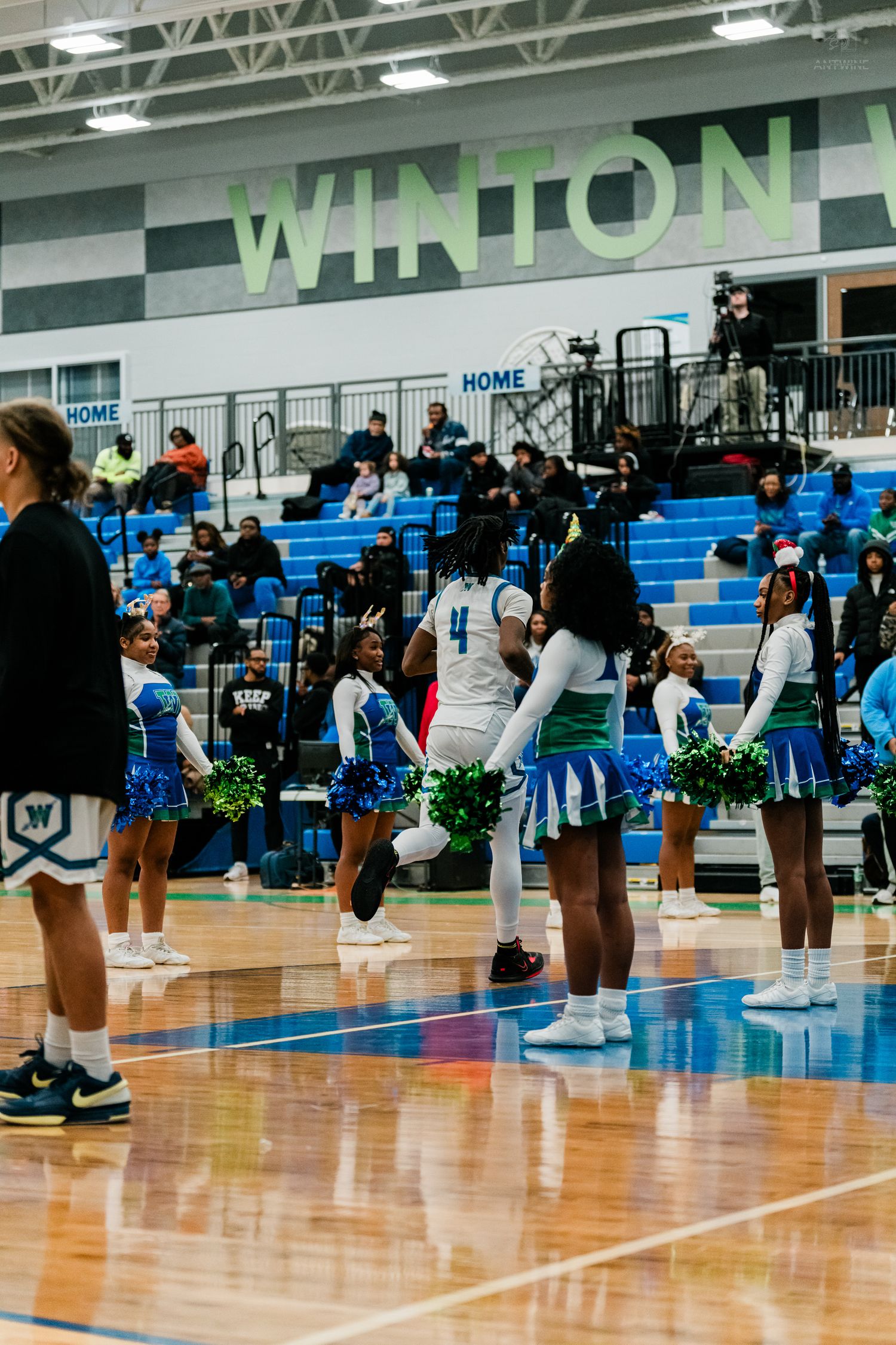 Basketball player #4 jogs past cheerleaders with green pom-poms during pregame at Winton school gymnasium.