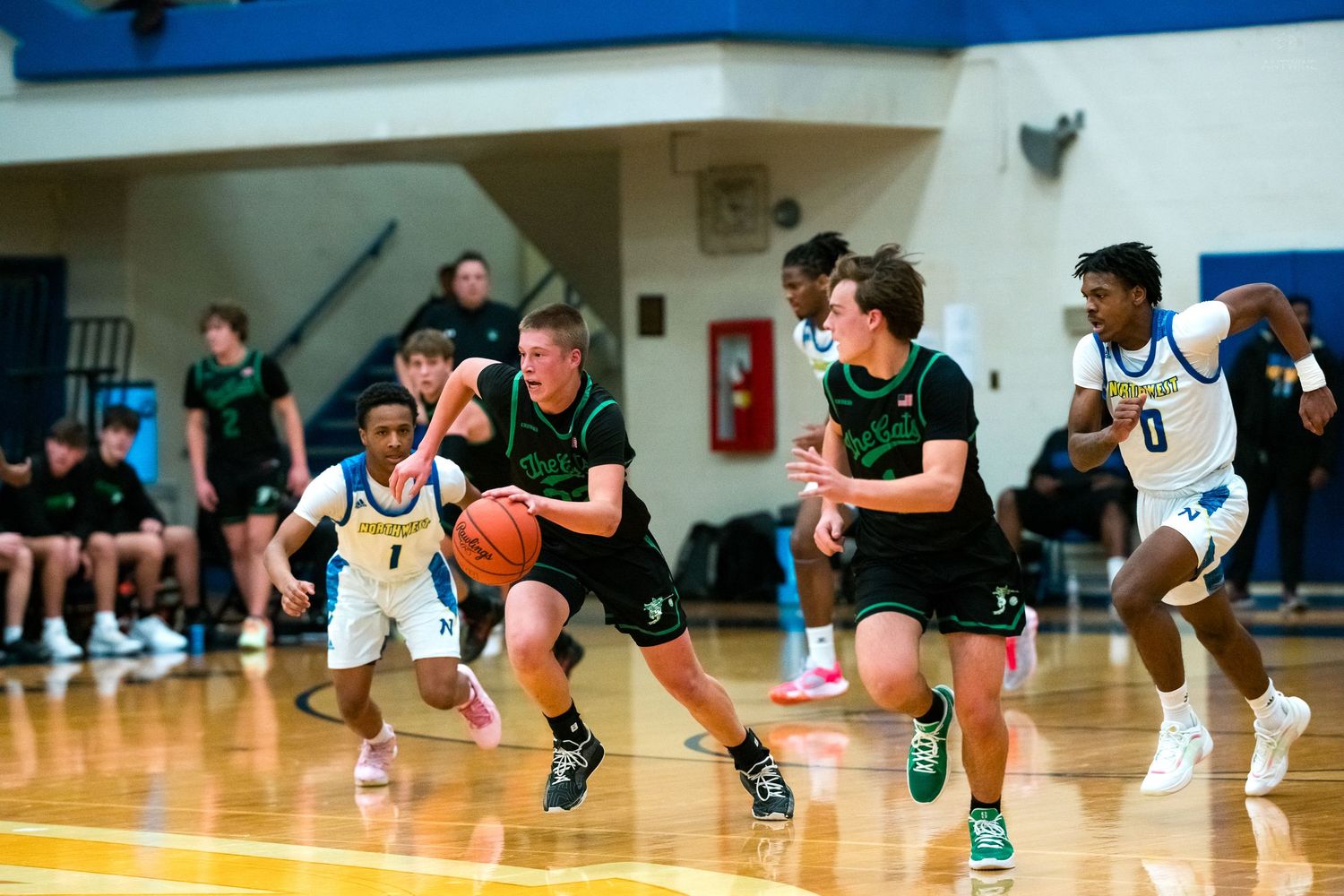 Green-uniformed basketball players drive downcourt during a competitive high school basketball game in a blue gymnasium.