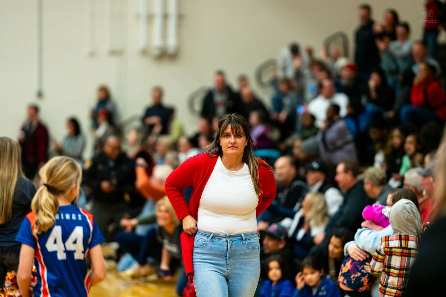A woman in a red cardigan walks on a basketball court sideline while a player wearing number 44 watches.