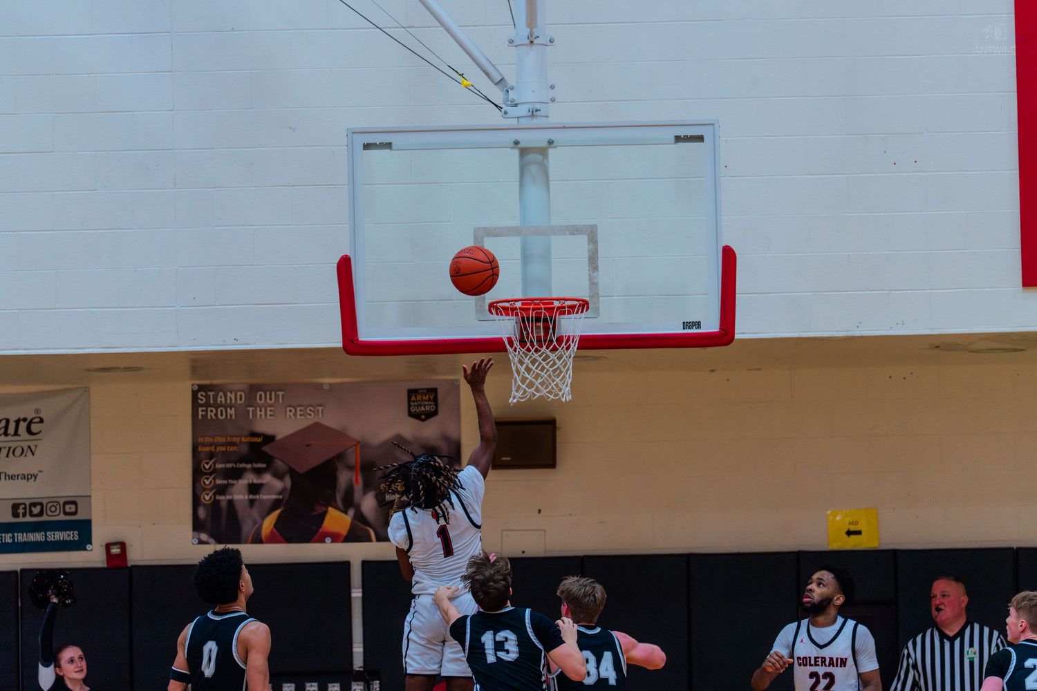 Basketball player scores layup while defenders in dark jerseys watch during high school gymnasium game.