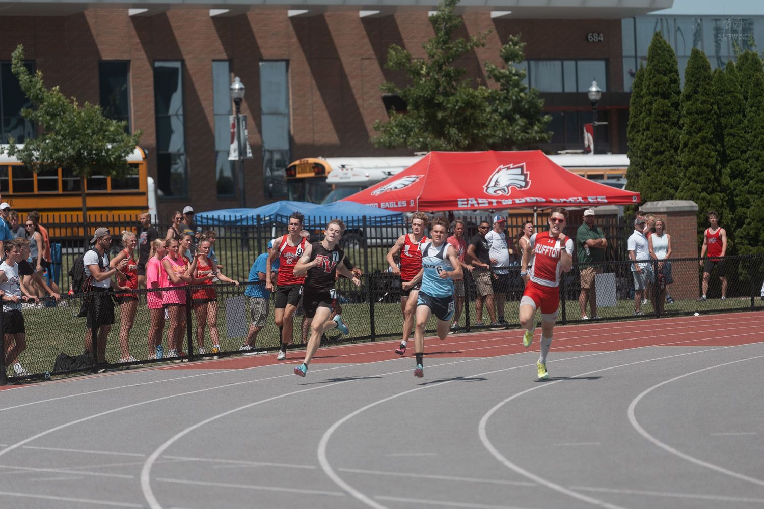 Competitors sprinting on a red outdoor track with cheering spectators during a high school track meet.