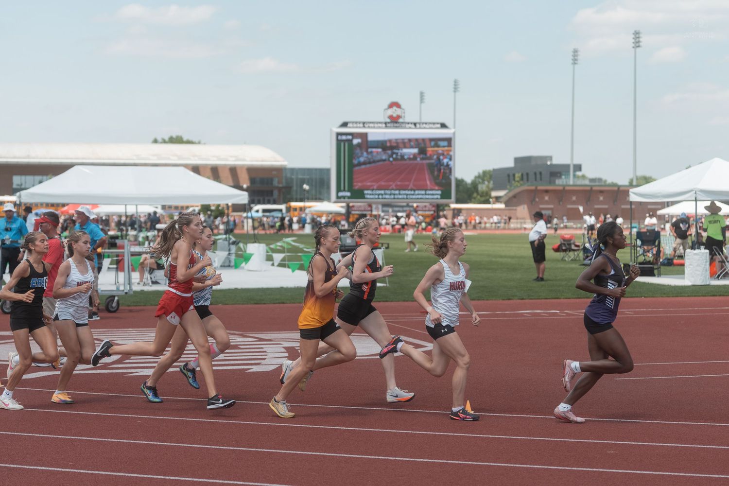 Male and female runners compete in a track race at an outdoor stadium with a scoreboard visible.